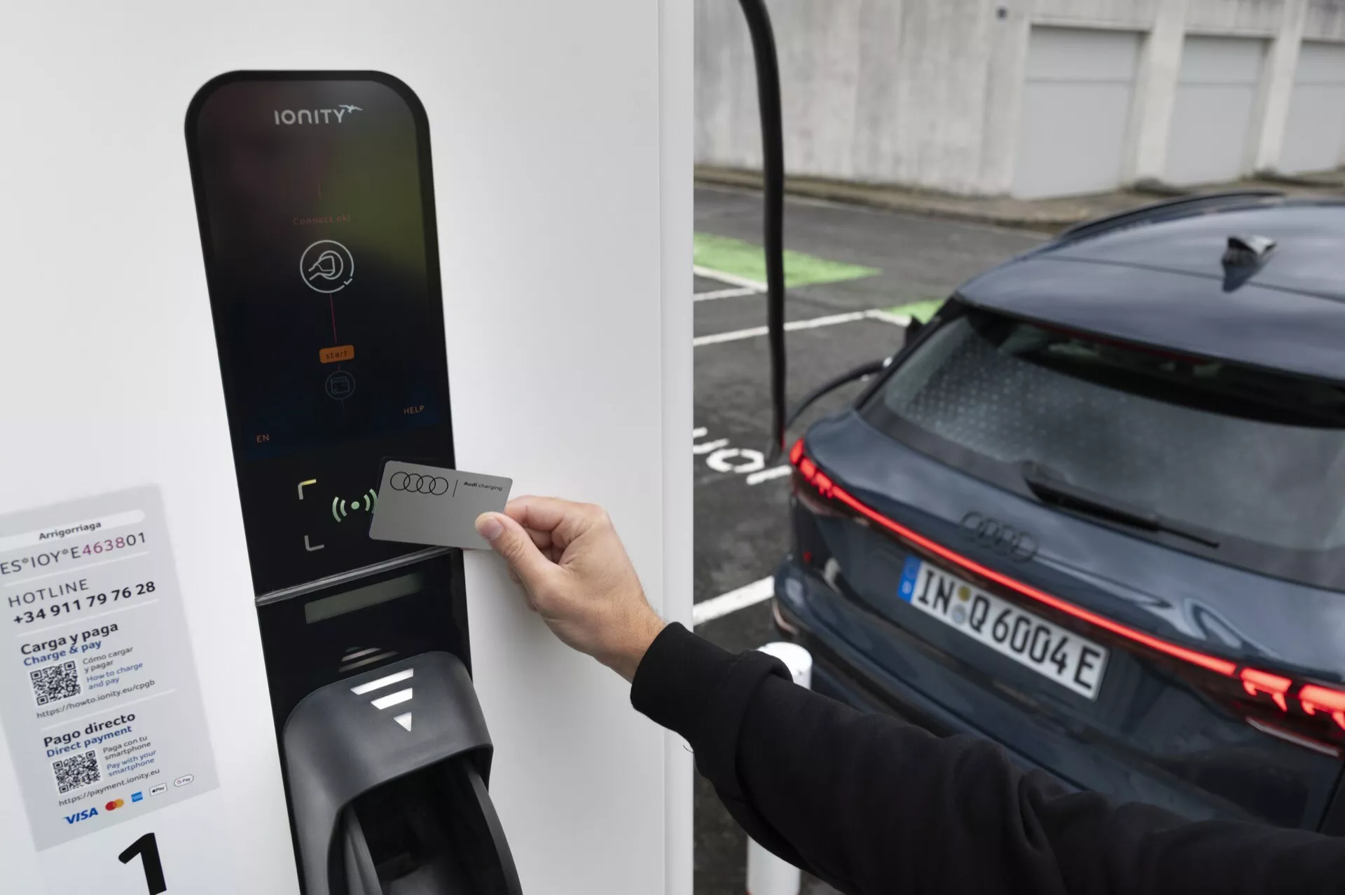 Obrázok osoby držiacej RFID kartu pri čítačke nabíjacej stanice pre elektromobily IONITY. Zamerané na zadnú časť moderného tmavého auta s horizontálnymi LED svetlami, čo naznačuje jeho elektrickú povahu. Auto, pravdepodobne EV, je zaparkované vedľa nabíjačky, čo naznačuje, že sa buď nabíja, alebo sa pripravuje na nabíjanie.