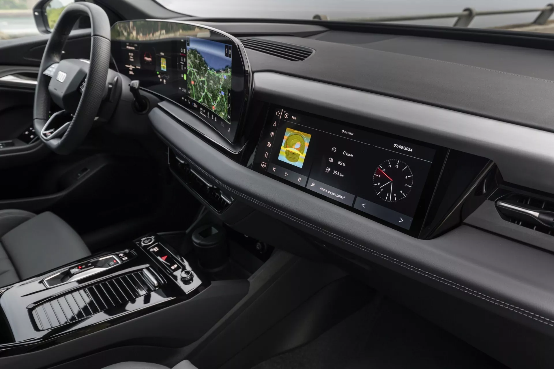 Pohľad na interiér Audi Q6 e-tron 2024 zameraný na palubnú dosku a centrálnu konzolu z mierne zvýšeného uhla. Vľavo je vidieť volant, čo naznačuje konfiguráciu na jazdu vľavo, zatiaľ čo centrálna konzola obsahuje radiacu páku, rôzne ovládacie prvky a veľký dotykový displej. Digitálny prístrojový panel zobrazuje jazdné metriky. Štýlová palubná doska sa horizontálne rozprestiera s vetracími otvormi a ďalším zobrazovacím prvkom vpravo. Pokročilá technológia je zrejmá z centrálneho dotykového displeja zobrazujúceho navigačné a multimediálne informácie, projekčného displeja viditeľného nad volantom a dotykových tlačidiel pre ovládanie klimatizácie. Dizajn zdôrazňuje minimalizmus a prémiové materiály, s koženým prešívaním a kovovými akcentmi, ktoré pridávajú na luxusnom pocite interiéru.