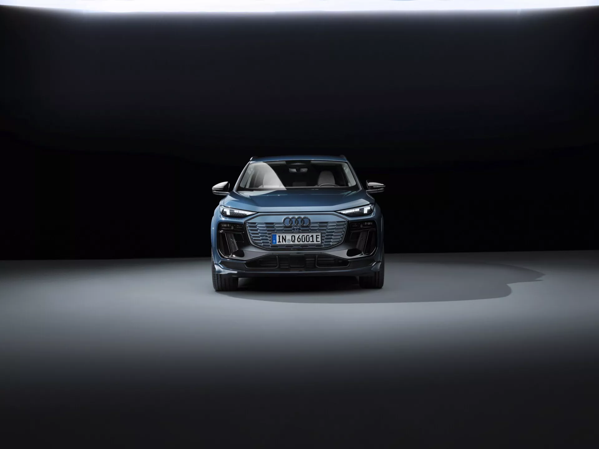 Pohľad spredu na Audi Q6 e-tron 2024, ktorý ukazuje kapotu, masku chladiča, časť strechy a predný dizajn. Elegantné svetlomety pravdepodobne disponujú LED technológiou. Nemecká poznávacia značka naznačuje elektrický alebo hybridný model. Výrazná maska Audi Singleframe, ďalšie otvory a možné bezpečnostné senzory, všetky zdôrazňujú jeho pokročilé asistenčné technológie. Dramatické osvetlenie s tmavým pozadím a osvetlenie zhora zvýrazňujú luxusný a sofistikovaný dizajn vozidla.