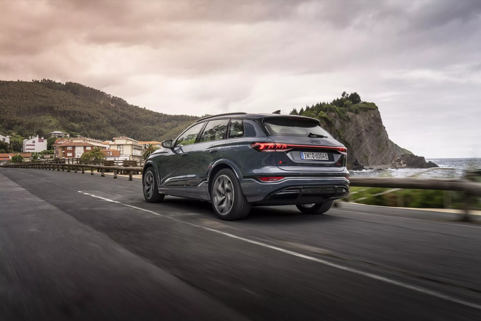 Audi Q6 e-tron 2024 zo zadného trojštvrťového pohľadu v pohybe na pobrežnej ceste, predvádzajúci výrazné LED zadné svetlá, tmavý metalický lak, zliatinové kolesá a praktický zadný stierač. Rozmazané pozadie naznačuje pobrežné prostredie, odrážajúc estetiku a technologický pokrok moderného SUV.