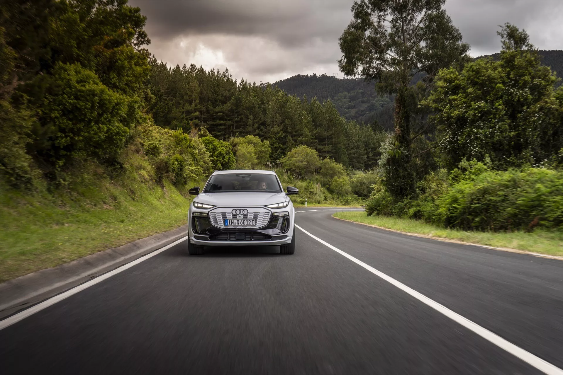 Audi Q6-e-tron 2024 zachytený v pohybe z predného uhla na dvojprúdovej ceste obklopenej zeleňou. Fotografia zobrazuje mriežku, svetlomety, kapotu a predný nárazník vozidla, zdôrazňuje jeho dynamický výkon a dizajn. Vozidlo má charakteristické LED svetlomety, výrazný luxusný znak na mriežke a aerodynamické línie, všetko za zamračeného počasia so suchými podmienkami na ceste.