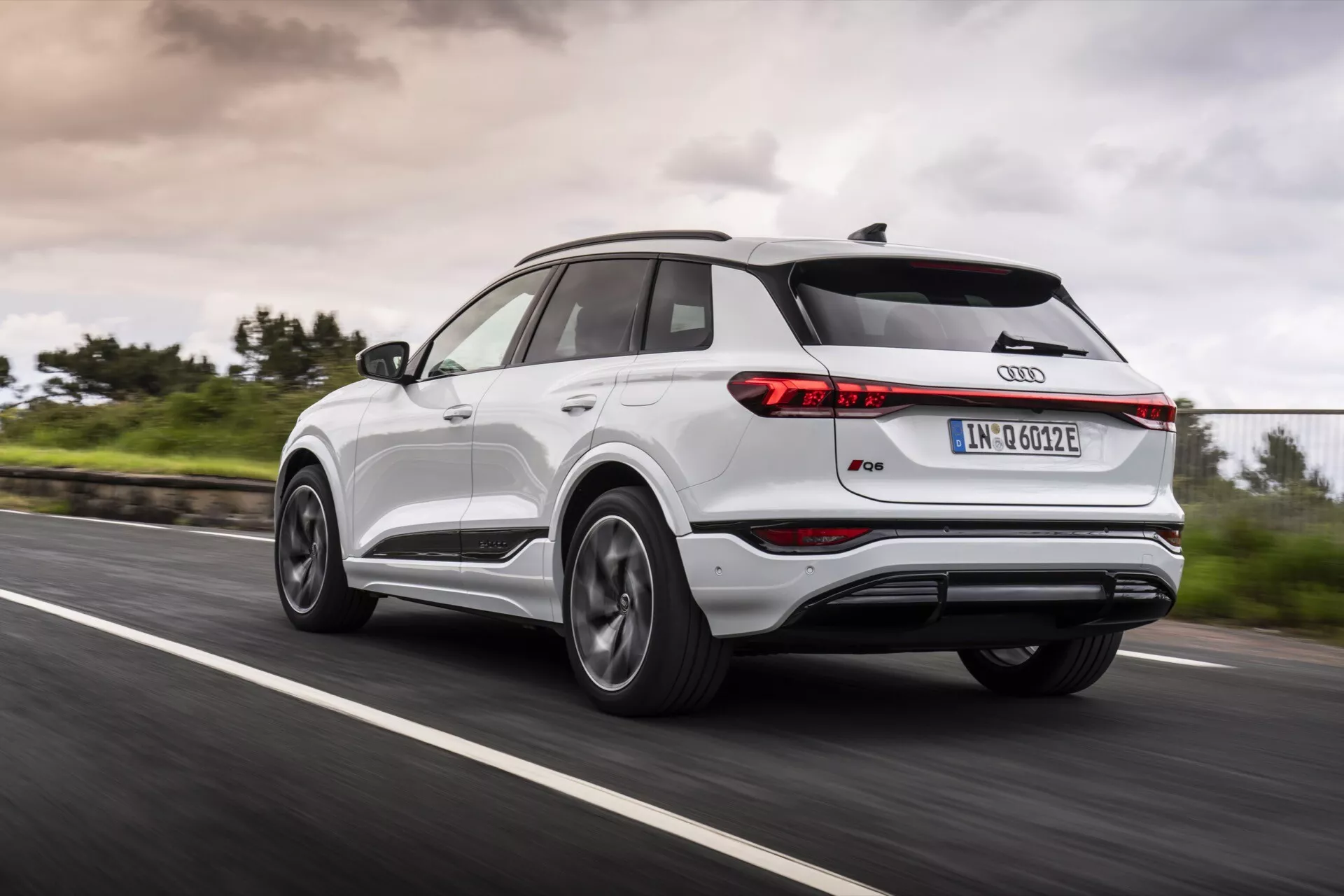 Audi Q4 e-tron 2024 idúce po asfaltovej ceste zobrazené z pohľadu zo zadnej tri štvrte, zachytávajúc ľavú zadnú stranu. Obrázok zobrazuje štíhle zadné svetlá, strešný spojler, panoramatickú strechu, kolesá z viacerými lúčmi z ľahkej zliatiny, dvojfarebnú karosériu s kovovými lištami a ikonickým znakom Audi. Rozmazanie pohybu kolies a zeleň pri krajnici s zamračenou oblohou naznačujú dynamické jazdné podmienky za akéhokoľvek počasia.