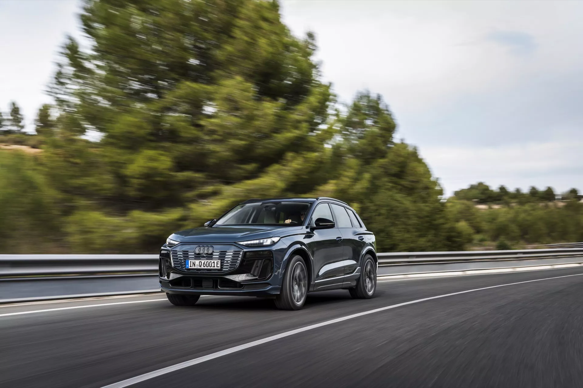 Audi Q6 e-tron 2024 SUV v pohybe na diaľnici, zachytený z trojštvrťového predného pohľadu. Obrázok zobrazuje prednú mriežku, LED svetlomety, kapotu, čelné sklo a časť bočného profilu, vrátane dverí a bočných zrkadiel. Vozidlo ide po pravej strane pruhu, pohybujúc sa zľava doprava s rozmazaným pozadím naznačujúcim rýchlosť. Stromy a zvodidlá sú mierne viditeľné. Číry dizajn predných svetlometov naznačuje pokročilé osvetľovacie systémy a veľká mriežka naznačuje technológie chladenia alebo senzory pre ADAS. Bočné zrkadlá môžu obsahovať smerové svetlá alebo systémy monitorovania mŕtveho uhla. Kolesá poskytujú pohľad do kategórií výkonnosti alebo luxusu.