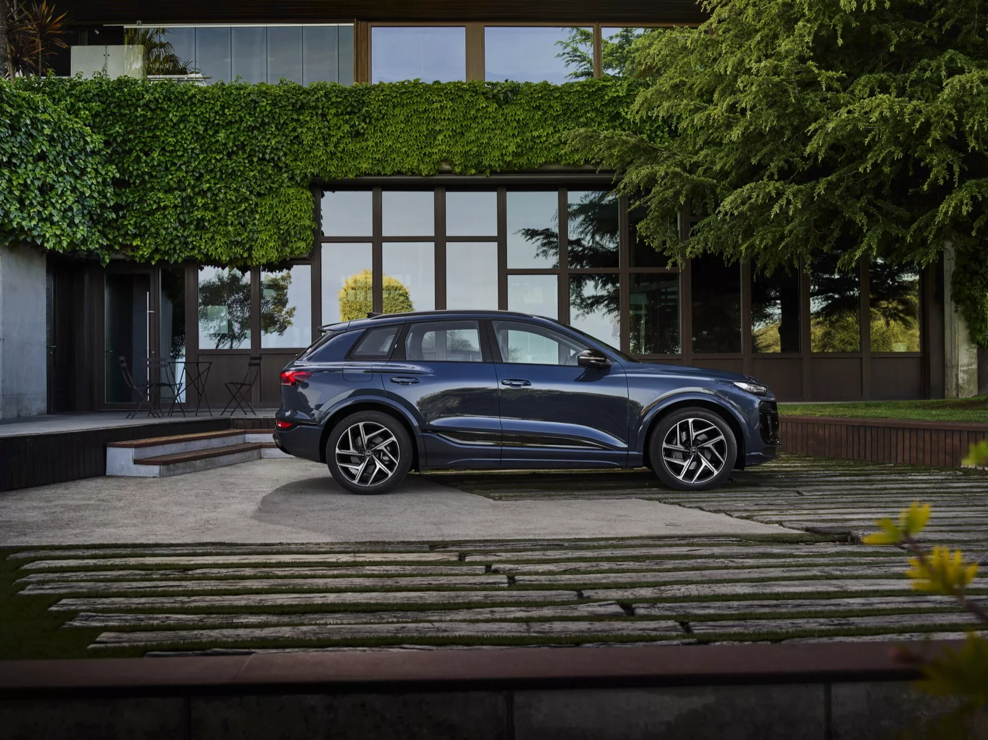 Audi Q6 e-tron 2024 stojí na dobre upravenej príjazdovej ceste, zachytené v kompletnom profilovom pohľade z mierne zvýšeného uhla. Hlbočizne kovové modré SUV sa leskne elegantným dizajnom, s aerodynamickými líniami strechy, tónovanými oknami a zapustenými kľučkami dverí. Viacpaprskové zliatinové kolesá a nízkoprofilové pneumatiky zdôrazňujú jeho špičkový výbavový stupeň. Na pozadí moderného luxusného budovy s veľkými sklenenými oknami a kamenami a trávnikovou príjazdovou cestou, je scéna jemne osvetlená, zvýrazňujúca prémiový status vozidla.