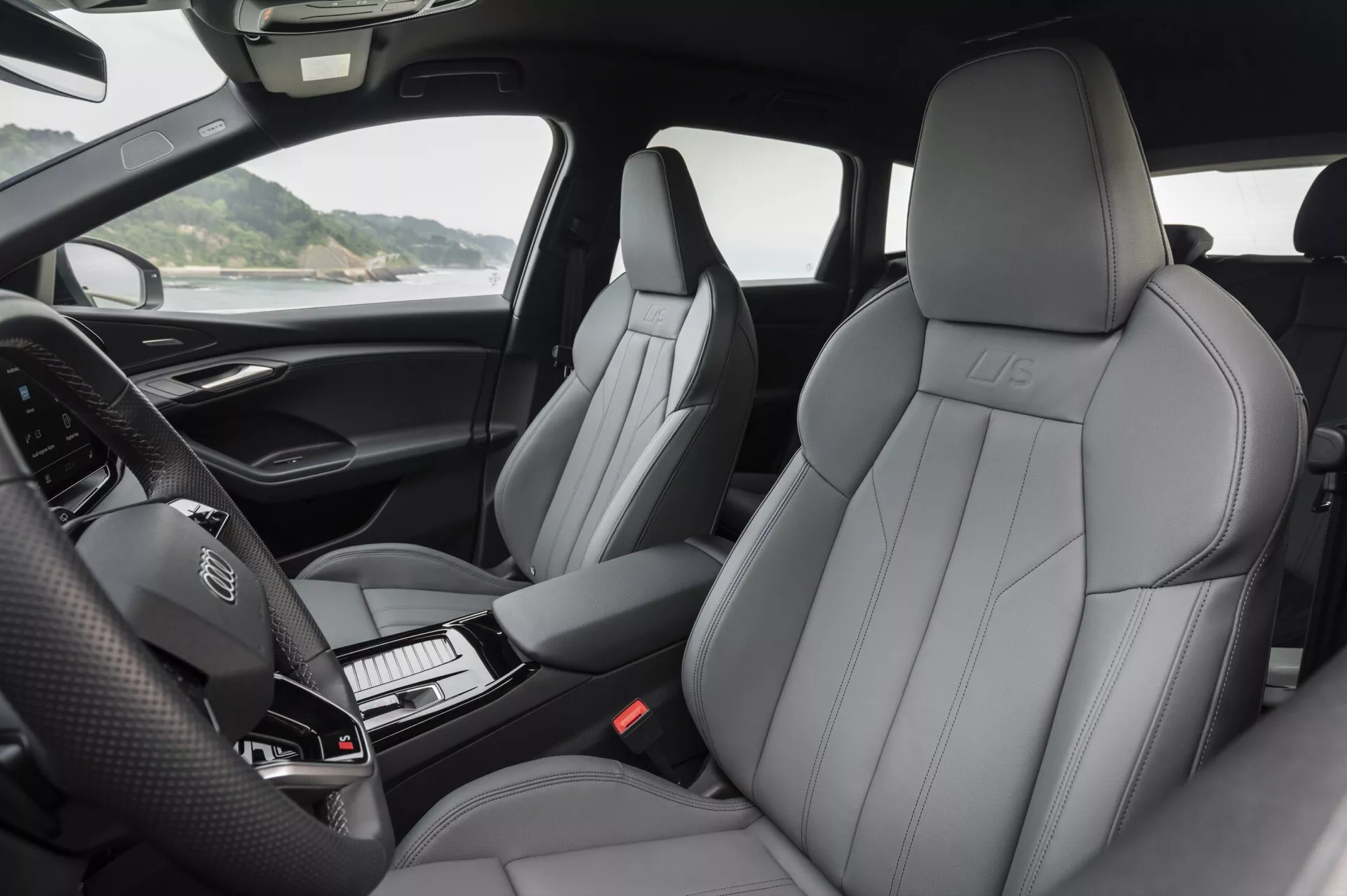 Pohľad na interiér Audi Q6 e-Tron 2024, ktorý ukazuje luxusné kožené predné sedadlá s výrazným šitím, embosovaným logom a moderným dizajnom. Fotografia zvýrazňuje prístrojovú dosku s volantom s integrovanými ovládacími prvkami a nezapnutým digitálnym prístrojovým panelom. Čiastočne je vidieť stredová konzola s radiacou pákou a usporiadanými tlačidlami. Obrázok zachytáva čalúnené opierky dverí s ovládaním pre zrkadlá, okná a pravdepodobne nastavenie sedadiel. Prirodzené svetlo zvýrazňuje komfort a vysokú kvalitu materiálov v kabíne.