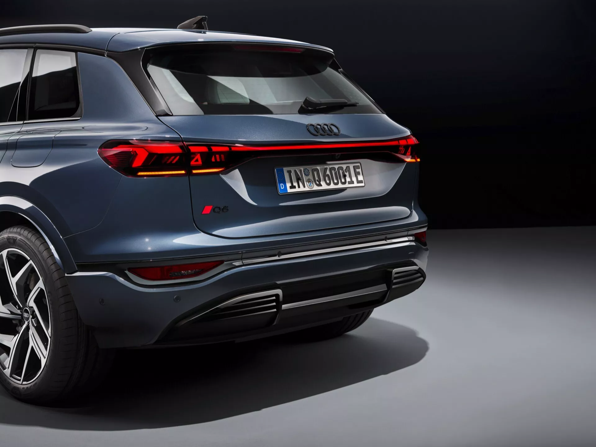Tento obrázok zobrazuje moderné SUV, pravdepodobne Audi Q6 e-tron 2024, z pohľadu zhora zozadu. Ikonické štyri kruhy Audi sú výrazne umiestnené vzadu, obklopené elegantným neprerušeným LED zadným svetlom, ktoré sa tiahne po celej šírke vozidla, so segmentovanými brzdovými svetlami a smerovkami. Tmavomodré auto sa pýši lešteným kovovým povrchom, odvážnym dizajnom nárazníka s integrovanými výfukmi a difúzorom, viacramennými zliatinovými kolesami s nízkoprofilovými pneumatikami a registračnou značkou naznačujúcou elektrický alebo hybridný pohon. Profesionálne osvetlenie a neutrálny pozadie zvýrazňuje luxusný a pokročilý dizajn vozidla, zdôrazňuje jeho športové a vysoké výkonné atribúty.