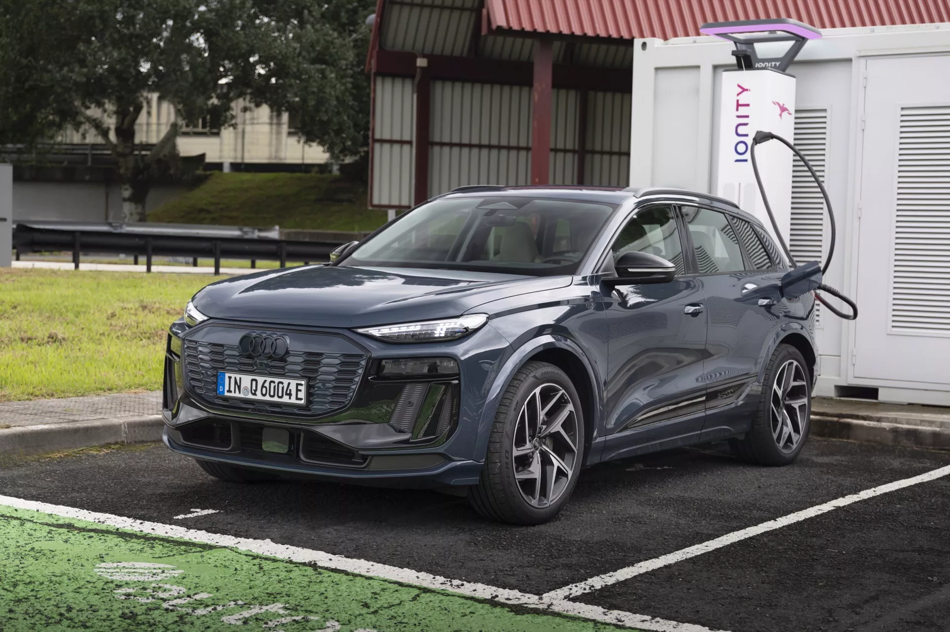 Obrázok ukazuje moderné SUV Audi Q6 e-Tron 2024 zaparkované pri nabíjacej stanici pre elektrické vozidlá. Auto je zobrazené z mierne šikmého predného pohľadu, čo zvýrazňuje jeho mriežku, svetlomety a časť kapoty a čelného skla. Je umiestnené diagonálne, pričom predná časť je mierne otočená doľava. Viditeľná je ľavá strana vozidla vrátane predného kolesa na strane vodiča, dvierok a bočného zrkadla. SUV je pripojené k nabíjacej stanici na ľavej prednej strane nad kolesom. Auto má elegantné línie, uzavretú mriežku bežnú pre elektrické vozidlá a pokročilé LED svetlomety. Detaily naznačujú možnosť pokročilých asistenčných systémov pre vodiča, ako je adaptívny tempomat, asistent udržiavania jazdného pruhu a systém prevencie kolízií. Obrázok je svetlý a čistý, vytvorený počas dňa bez pohybového rozmazania, takže všetky dizajnové a technologické prvky sú ľahko viditeľné.