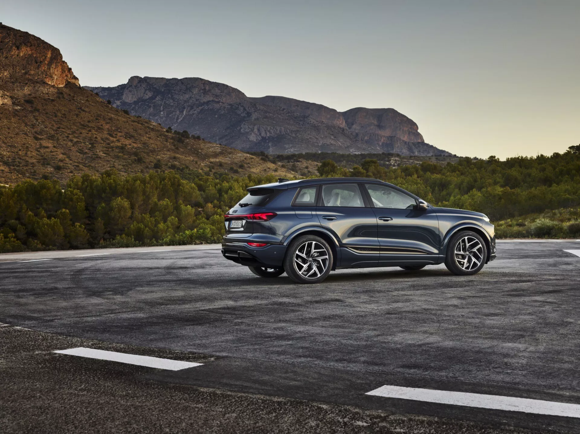Obrázok SUV Audi Q6-e-tron 2024 zaparkovaného na vonkajšom parkovisku s prírodnou, možno horskou krajinou v pozadí. Fotografia zachytáva trojštvrtinový zadný pohľad, ktorý zvýrazňuje bočné aj zadné dizajnové prvky. Strana vodiča je zreteľne viditeľná, ukazujúc dizajnové línie, zadné okná a dizajn kolies. Sklonená línia strechy naznačuje športový, kupé-SUV dizajn. Moderné zadné LED svetlá, viditeľné senzory alebo odrazky na zadnom nárazníku a dvojfarebná farebná schéma zdôrazňujú výšku a postoj. Veľké hliníkové kolesá a výrazné pneumatiky naznačujú vyšší výbavový stupeň so športovým vzhľadom. Vozidlo sa zdá byť blízko kameře, poskytujúc detailný pohľad. Významné prvky zahŕňajú LED osvetlenie, parkovacie senzory, vysokovýkonné kolesá a pneumatiky, aerodynamický dizajn a možne pokročilé asistenčné systémy pre vedenie a pohon všetkých štyroch kolies.