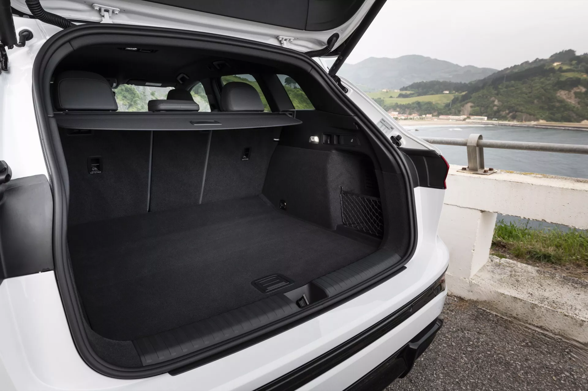 Obrázok znázorňuje nákladný priestor bieleho Audi Q6 e-Tron 2024 hatchbacku alebo SUV, zobrazený zozadu s otvoreným vekom kufra a miernym sklopením kamery nadol. Kufor s čiernym kobercom je prázdny, čo ukazuje jeho priestrannosť a čistotu. Zadné sedadlá sú vzpriamené, čo naznačuje možnosť prepravy cestujúcich alebo sklopenia sedadiel pre väčší nákladový priestor. Zakrytí nákladu sa nachádza hneď za zadnými sedadlami, ale nie je rozložené, čo umožňuje úplný pohľad do kufra. Na ľavej strane nákladného priestoru je 12-voltová zásuvka a na pravej strane sa zdá byť odkladací priestor pre uloženie drobných predmetov. Viditeľný je zadný stierač. Vozidlo je zaparkované pri pobreží, čo naznačuje prímorskú lokáciu. V kufri nie sú žiadne predmety ani ľudia.