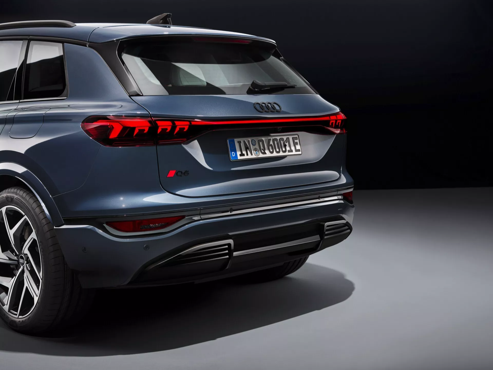 Pohľad zozadu na Audi Q6-e-tron 2024 zobrazujúci dizajn zadných svetiel s LED technológiou, výrazný spojler na veku kufra, zadný nárazník s difúzormi a senzormi pre ADAS, európsky evidenčný štítok a čiastočne viditeľné označenie modelu naznačujúce elektrické alebo plug-in hybridné SUV z Audi Q-série. Štúdiové prostredie s reflexnou leštenou podlahou.