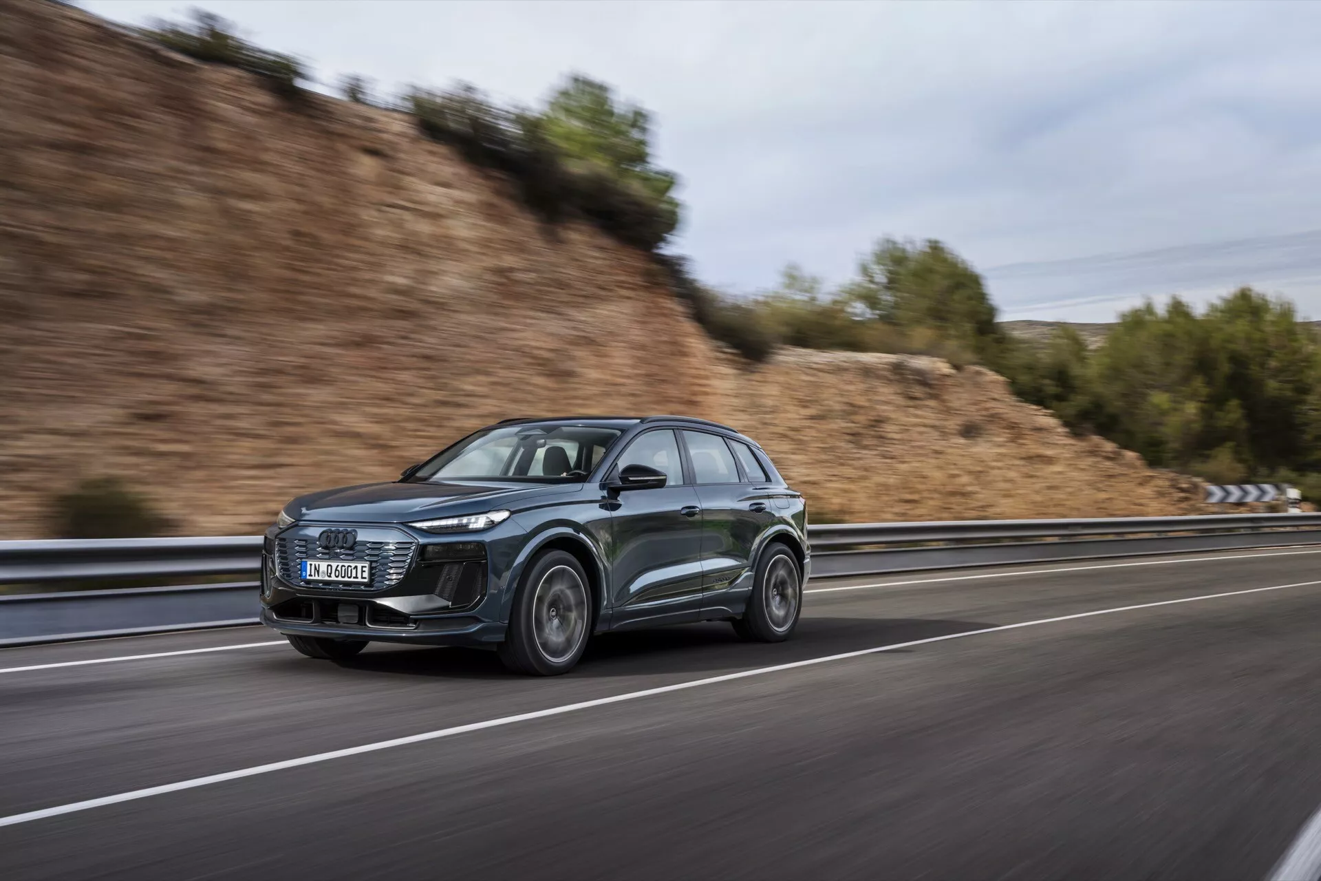 Tento obrázok zobrazuje moderné a sofistikované Audi Q6 e-tron 2024 SUV, fotografované z bočného uhla pri pohybe. Moderný dizajn vozidla je zvýraznený výraznými LED svetlometmi a aerodynamickými prvkami, vrátane šikmej strechy a aerodynamických bočných zrkadiel. Auto, dokončené v tmavej farbe, pravdepodobne šedej alebo čiernej, ide po rýchlostnej ceste, čo je vidieť na rozmazanom pozadí vytvorenom pomocou techniky panoramatickej fotografie. Predné koleso je mierne otočené smerom ku kamere, čo ukazuje dizajn zliatinových kolies. Ďalšie viditeľné prvky zahŕňajú možnú integrovanú technológiu v svetlometoch a bočných zrkadlách, ako aj možné strešné lišty zamerané na úžitkovosť.