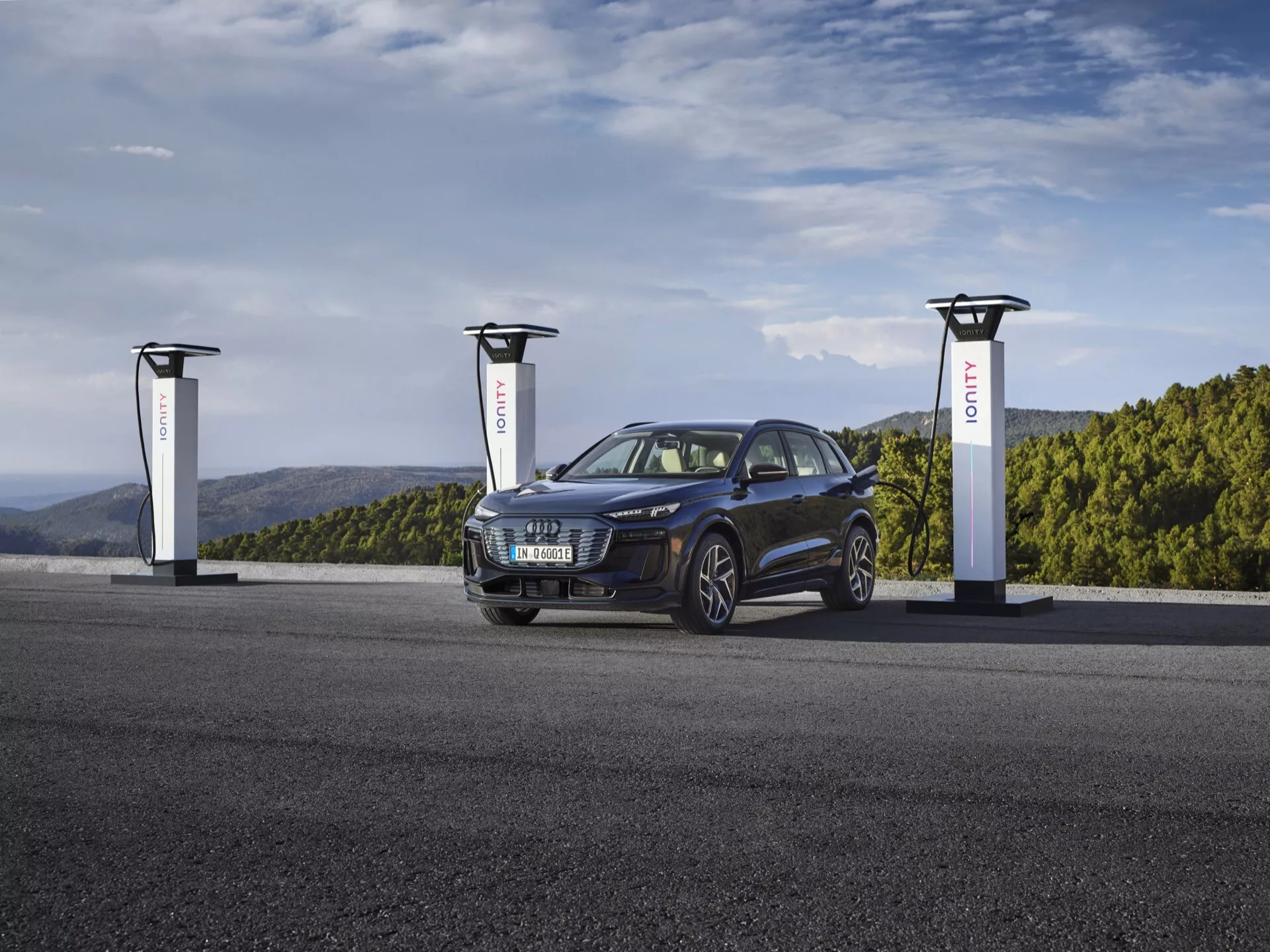 SUV Audi Q6 e-tron 2024 v tmavomodrej metalíze na nabíjacej stanici pre elektromobily s tromi nabíjačkami. Obrázok zachytáva vozidlo v uhle troch štvrtín, ukazujúc jeho prednú ľavú stranu, zvýrazňujúc mriežku, svetlomety, kolesá a kapotu. Štýlové a moderné elektrické vozidlo má športový a všestranný dizajn s elegantnými, agresívnymi svetlometmi a zliatinovými kolesami, zdôrazňujúc pokročilú a udržateľnú automobilovú technológiu.