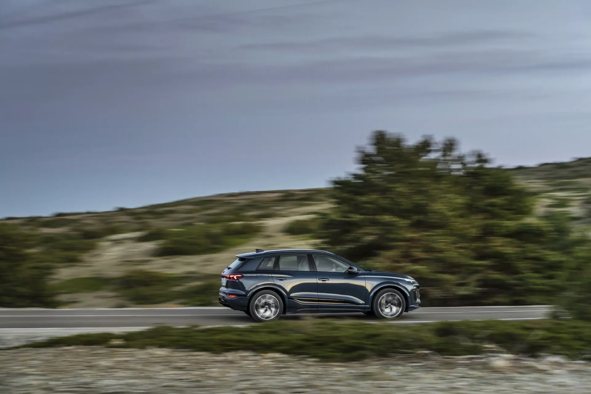 Dynamický bočný pohľad na Audi Q6 e-tron 2024 v pohybe s rozmazaným pozadím, stredný paralelný uhol zobrazujúci profil, dizajnové línie, okná, päť dverí, bočné zrkadlá, hliníkové ráfiky, denné nastavenie, vidiecke pozadie.