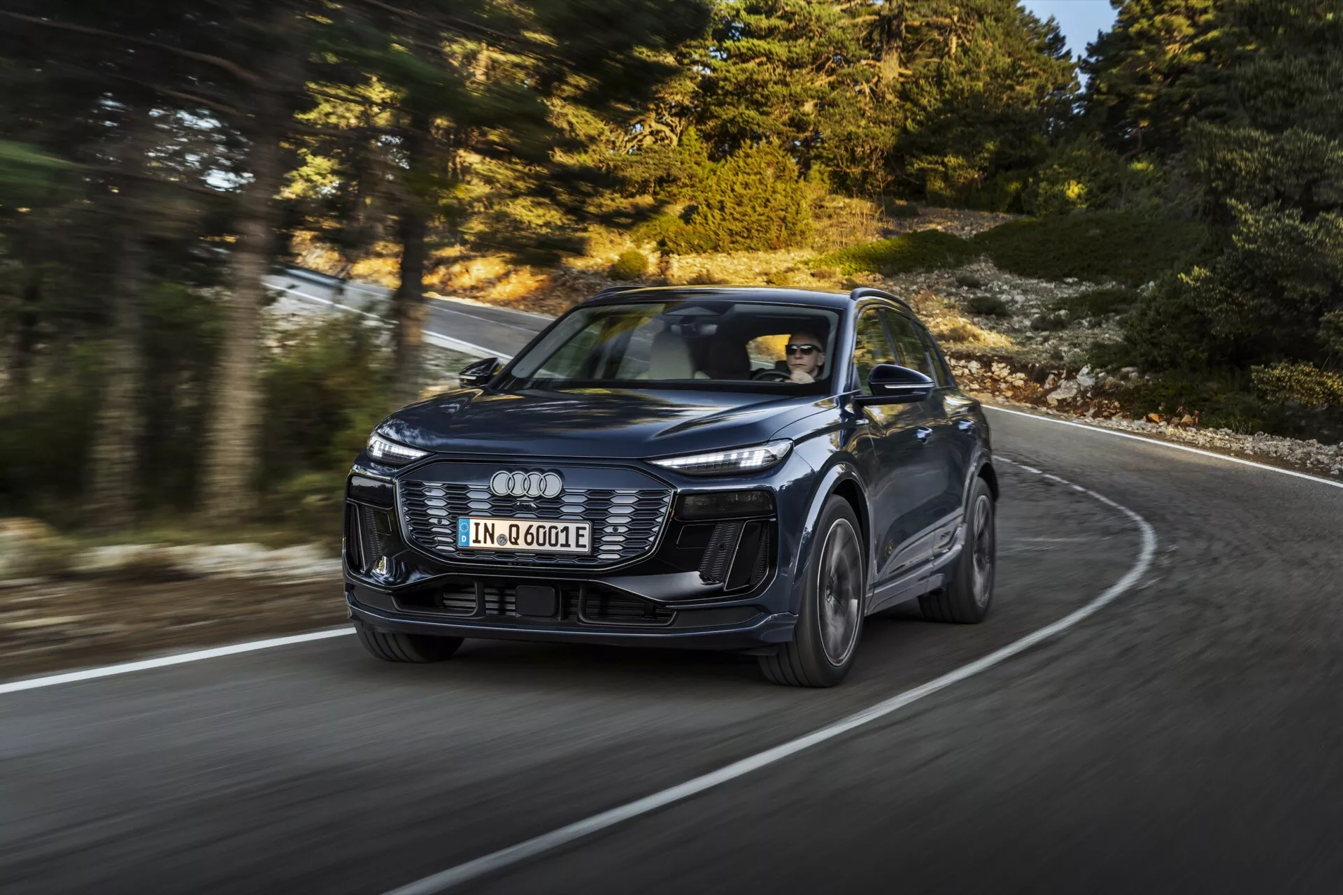 Obrázok moderného SUV Audi Q6 e-tron 2024 v pohybe, zachytený z dynamického trojštvrťového predného uhla, zvýrazňujúci dizajn a výkon. Vozidlo, identifikované podľa charakteristického loga so štyrmi kruhmi, má emblémové LED svetlá, agresívny predný nárazník a tvarované línie kapoty. Obrázok, urobený z nižšieho uhla, zvýrazňuje prítomnosť vozidla na zakrivenej ceste, pričom jeho zliatinové kolesá sú mierne rozmazané, čo naznačuje pohyb. Nový model naznačuje pokročilé technológie, ako je adaptívny tempomat a elektrický pohon, označené odznakom 'e-tron'. Profesionálne fotografovanie, ktoré predstavuje luxus, dizajn a technológiu v konfigurácii s ľavostranným riadením.