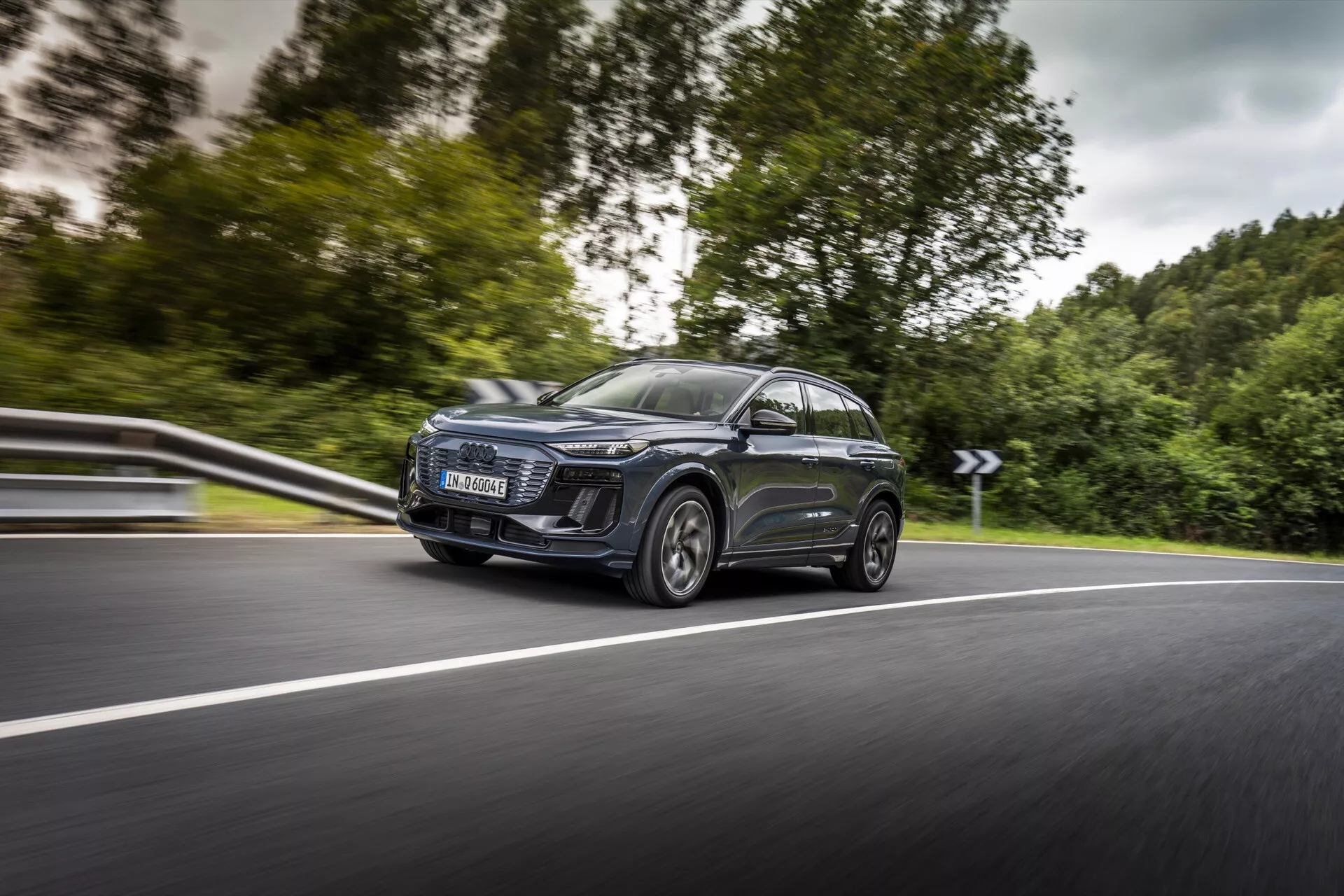 Na obrázku je moderné SUV Audi Q6 e-tron 2024 v pohybe, zachytené z uhla predného boku, so zameraním na jeho ľavú stranu. Dynamický záber zdôrazňuje výrazný predný ľavý svetlomet s výrazným LED denným svietením, veľkú modernú mriežku, štýlové aerodynamické bočné zrkadlá s integrovanými smerovými svetlami a veľké zliatinové kolesá s nízkoprofilovými pneumatikami. Auto má panoramatickú strechu a tónované okná, so zrkadlovým kovovým lakom. Nízky uhol pohľadu a mierne rozmazané pozadie zdôrazňujú rýchlosť a pohyb auta na zakrivenej dvojprúdovej ceste.