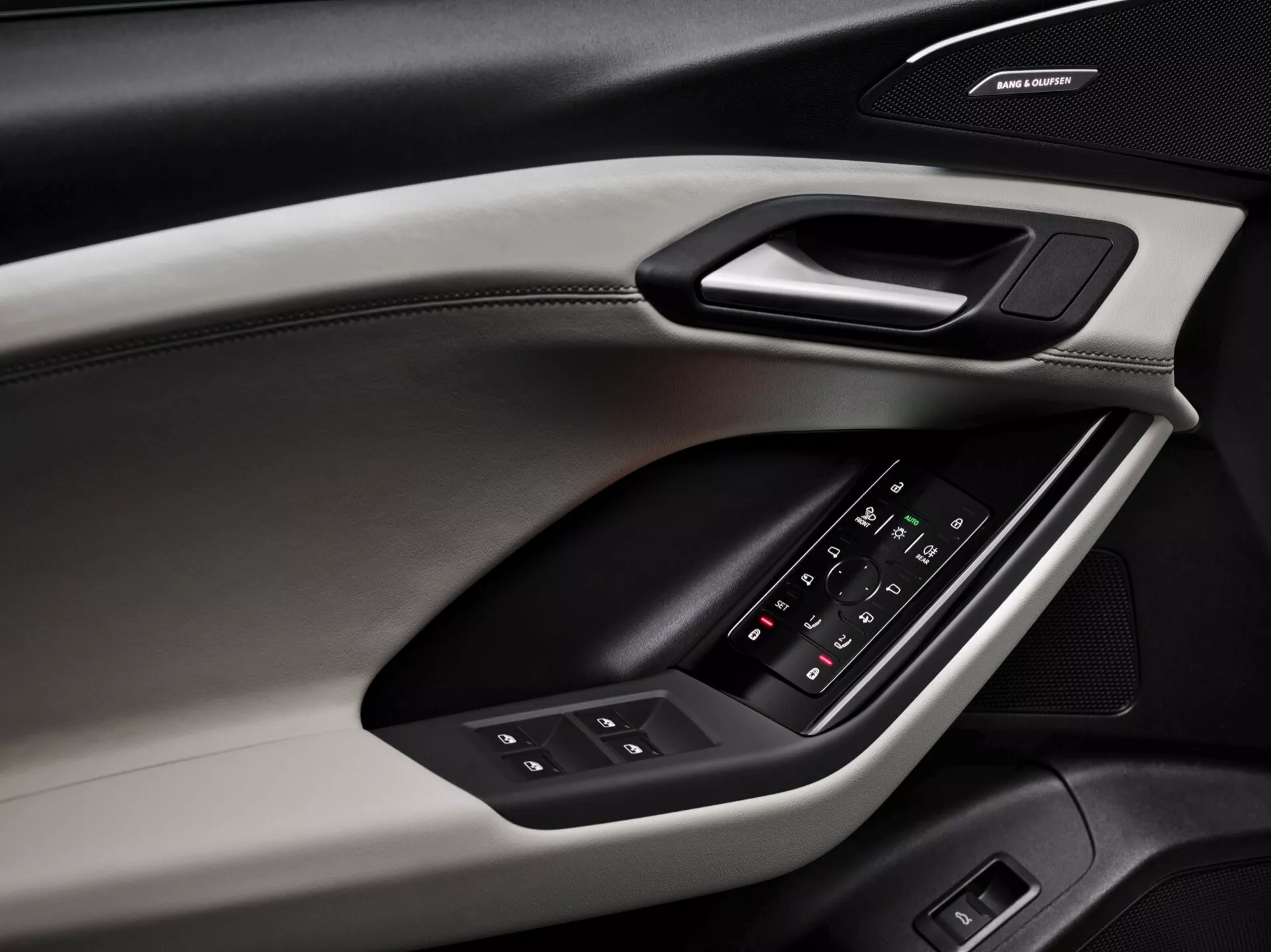 Detailný pohľad na vnútorný dverový panel Audi Q6 e-tron 2024, zdôrazňujúci dizajnové prvky a funkcie. Dverový panel, snímaný z uhlopriečneho uhla, odhaľuje rôzne materiály a textúry, vrátane mäkkej hornej časti s viditeľným prešívaním, tvrdého obloženia obsahujúceho ergonomickú kľučku a polstrovanú lakťovú opierku s podsvieteným ovládacím panelom pre okná a nastavenie spätných zrkadiel. Pod lakťovou opierkou je značkový reproduktor, ktorý naznačuje prémiový audio systém. Farebná schéma zahŕňa dvojfarebný dizajn, ktorý zvyšuje estetické a funkčné vlastnosti.