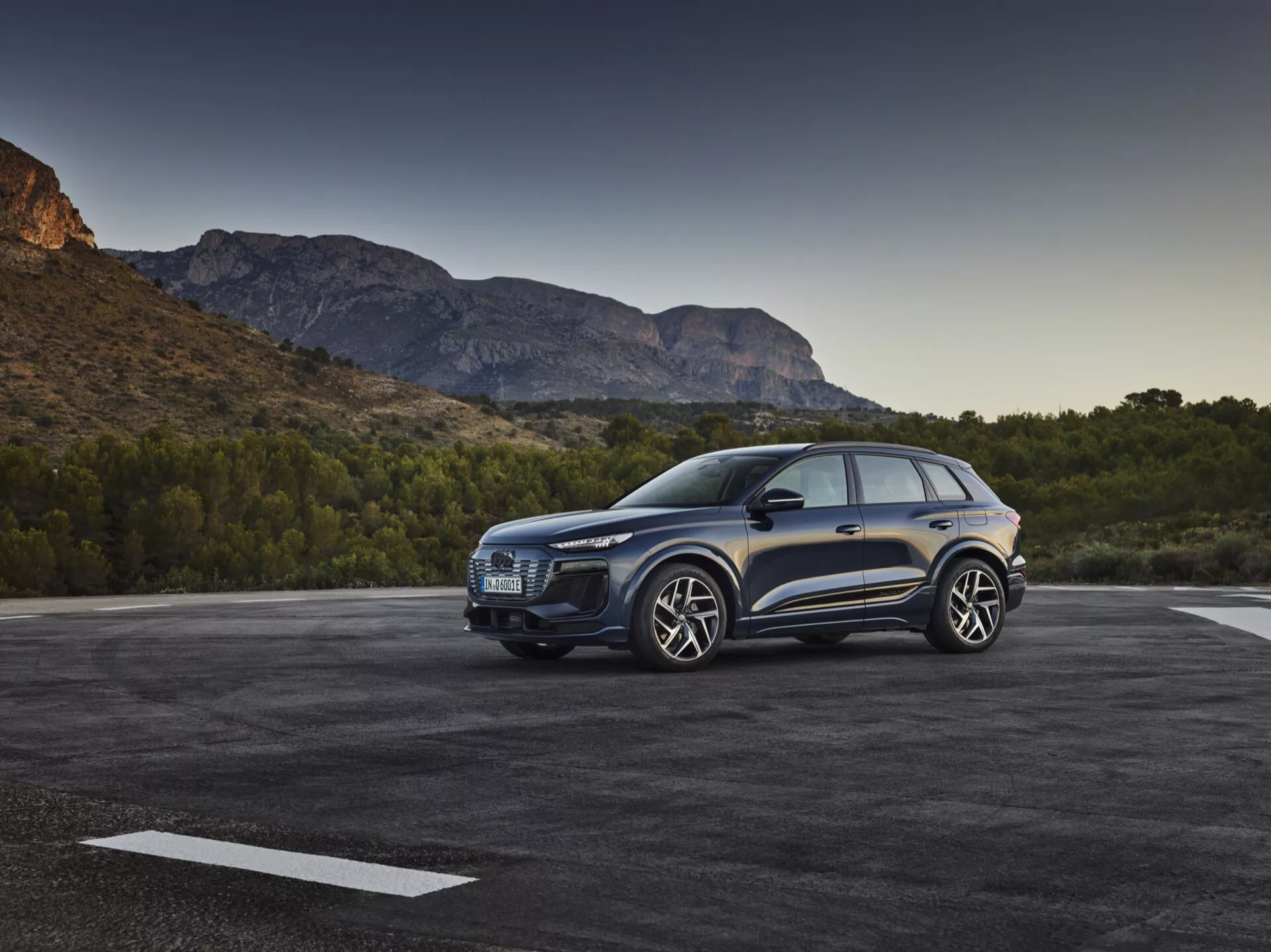 Trojštvrťový predný pohľad na modré SUV Audi Q6 e-tron 2024 zaparkované vonku, ukazujúci výraznú mriežku, elegantné svetlomety, tvarovanú kapotu, veľké zliatinové kolesá, bočné zrkadlá a dvere vodiča na pozadí malebného horského prostredia pri rannom alebo neskorom popoludňajšom svetle.