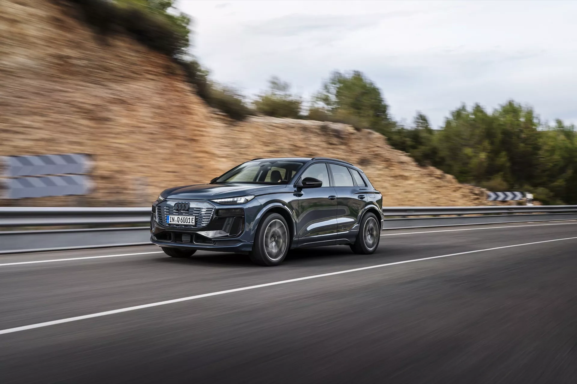 Obrázok Audi Q6 e-tron 2024 v pohybe, vytvorený pomocou techniky panningu, ktorá zdôrazňuje elegantný profil a moderný dizajn SUV. Auto, rámované zo strany, sa rýchlo pohybuje po viacprúdovom diaľničnom pruhu s rozmazanými kolesami a pozadím, čo podčiarkuje jeho dynamické schopnosti. Tmavý kovový voz má veľkú prednú mriežku, výrazné LED svetlá, športové zliatinové kolesá a reflexné povrchy. Zamračené nebo a mokrá cesta vytvárajú dramatickú scénu, zvýrazňujúc luxusné a výkonné vlastnosti auta.