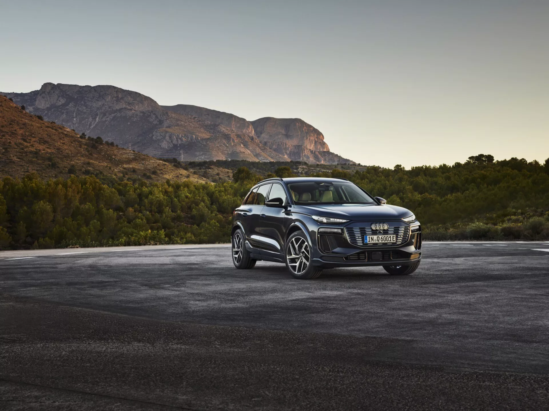 Luxusné SUV Audi Q6 e-tron 2024 v prednom trojštvrťovom uhle, zobrazujúce chromovanú mriežku, moderné LED svetlomety, veľké viaclúčové zliatinové kolesá, bočné zrkadlá s integrovanými smerovkami, tónované okná a lesklý lak, zaparkované na asfalte s horami v pozadí za úsvitu alebo súmraku.
