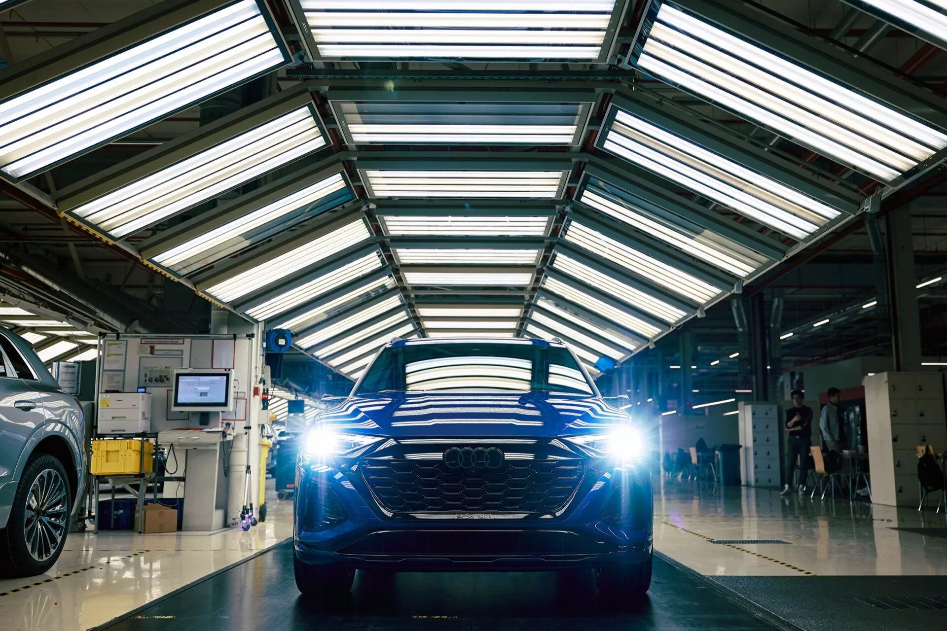 Pohľad spredu na Audi Q8 e-tron 2023 v dielni s odrazivou podlahou, zdôrazňujúci jeho horizontálnu mriežku a LED svetlomety.
