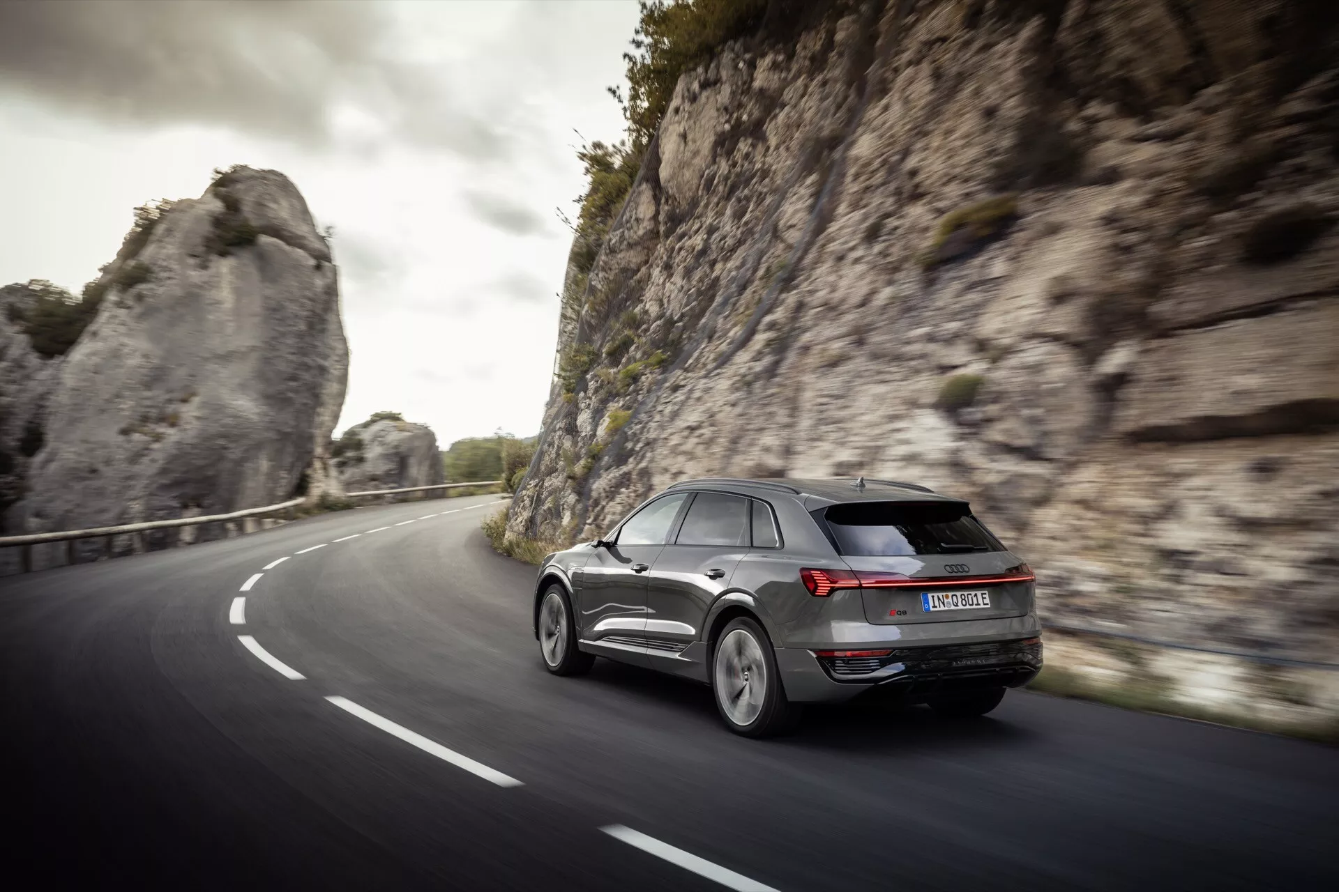 Moderné SUV Audi Q8 e-tron 2023 v dynamickom zadnom trojštvrťovom pohľade idúce po zakrivenej dvojprúdovej ceste v prírodnom, možno hornatom prostredí, predvádzajúce LED zadné svetlá, zliatinové kolesá a aerodynamický dizajn v mäkkom osvetlení