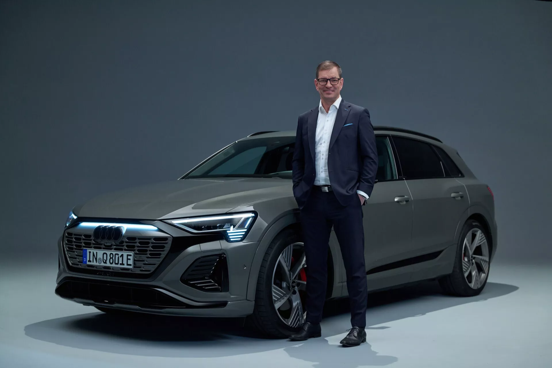 Audi Q8 e-tron 2023 v štúdiovom prostredí, predstavujúce prednú mriežku, LED svetlomety, viaceramenné zliatinové kolesá a elegantný štýl karosérie SUV s mužom v obleku pre mierku