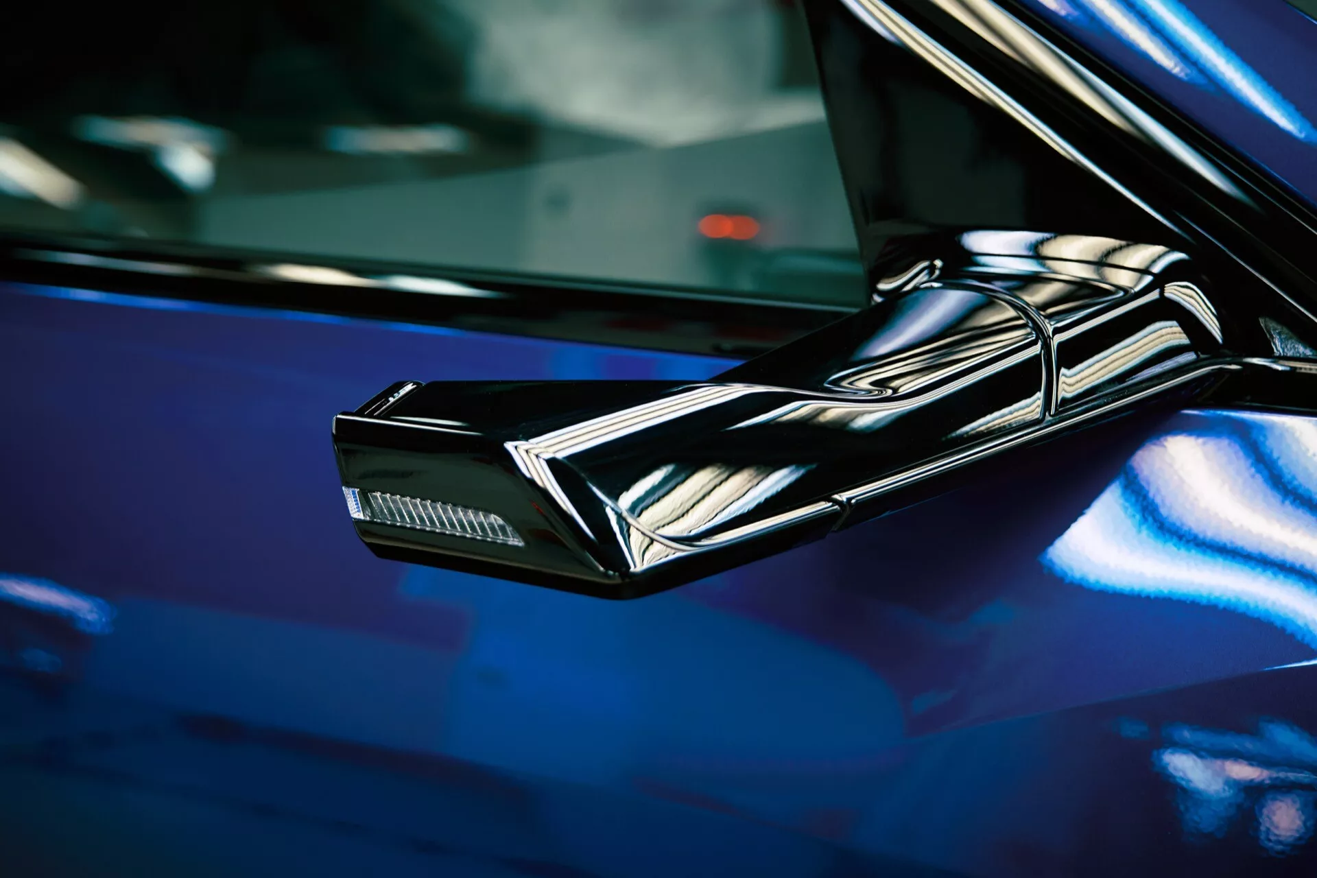 Detailný pohľad na elegantné, uhlové, čierne bočné zrkadlo Audi Q8 e-tron 2023, ktoré zdôrazňuje futuristické dizajnové prvky a integrované smerové svetlá na pozadí hlboko modrej, lesklej povrchu auta