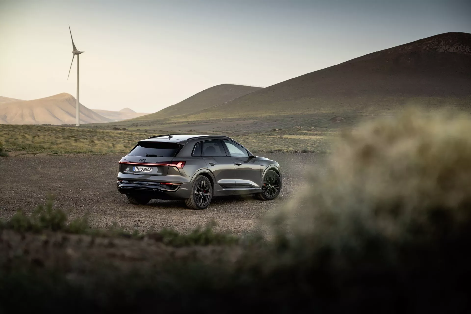 Audi Q8 e-tron 2023 v prírodnom vonkajšom prostredí s veternými turbínami na pozadí, prezentujúci jeho športový dizajn s panoramatickou strechou, duálnymi výfukmi a trojštvrťovým pohľadom zozadu v neskoré popoludnie
