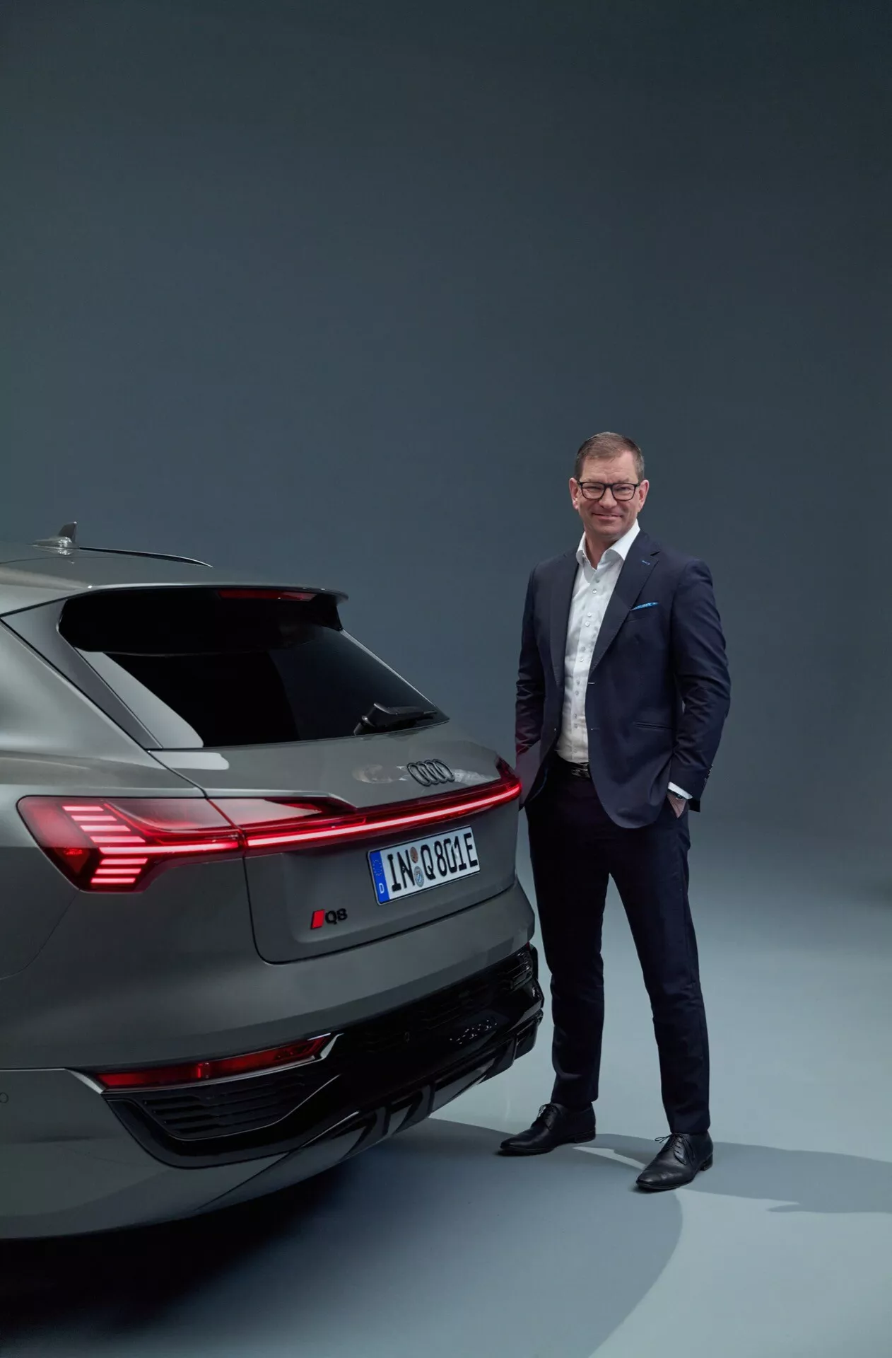 Elektrické vozidlo Audi Q8 e-tron 2023 ukazujúce zadný dizajn vrátane zadných svetiel, nárazníka a výfukového systému v ateliérovom nastavení, s profesionálne oblečeným mužom stojacim vedľa, zdôrazňujúce modernú automobilovú technológiu a luxus.
