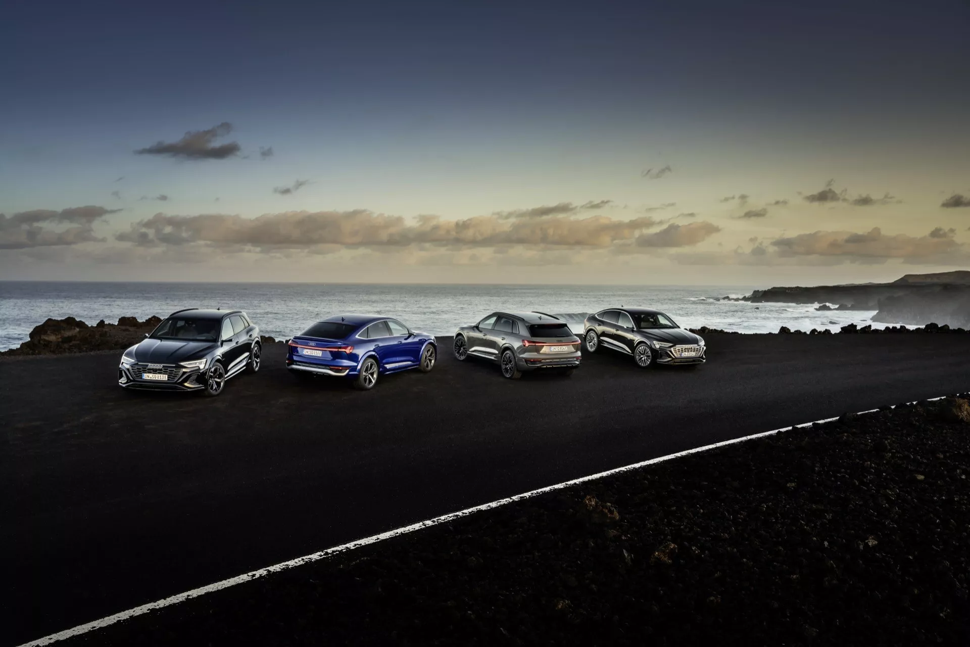 Audi Q8 e-tron 2023 zaparkované na asfaltovej ceste v pobrežnom prostredí so skalnatým terénom a výhľadom na oceán v súmraku, ukazujúce aerodynamický dizajn, zliatinové kolesá a moderné osvetlenie