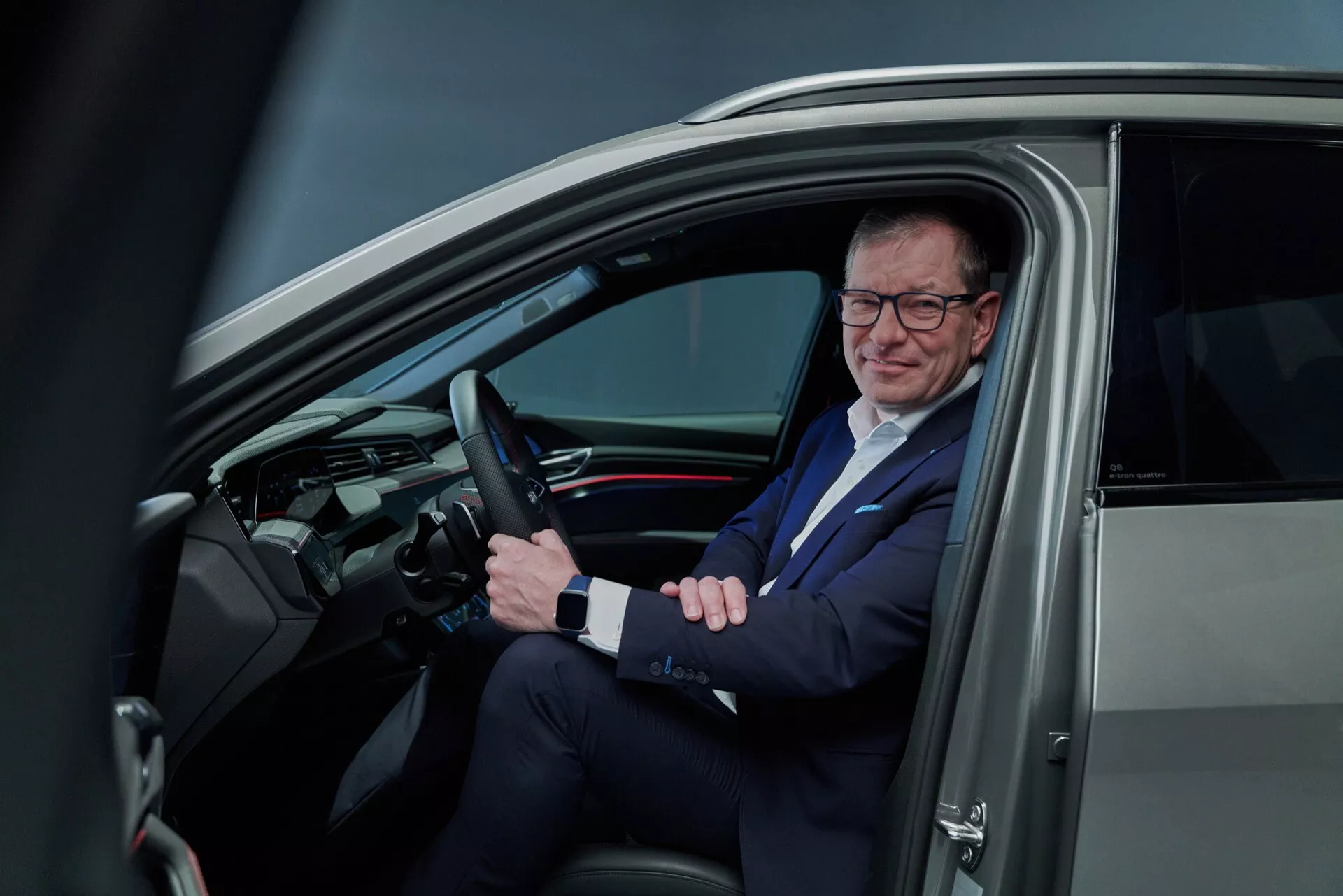 Pohľad do interiéru Audi Q8 e-tron 2023 cez otvorené dvere vodiča ukazujúci volant s ovládacími prvkami, elegantnú palubnú dosku s digitálnym displejom a muža v obleku na sedadle vodiča, čo odráža modernú technológiu vozidiel a podnikateľský životný štýl