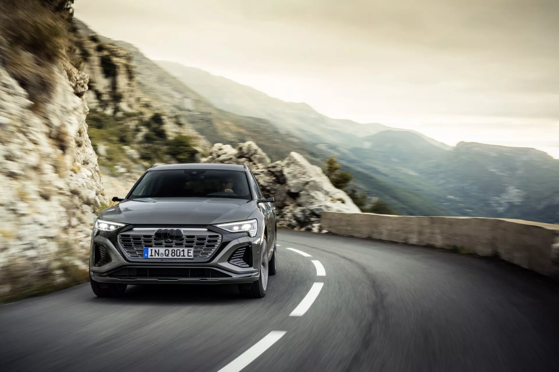 Audi Q8 e-tron 2023 na zákrute ceste so skalnatými horami v pozadí, predvádzajúce aerodynamický dizajn a pokročilú technológiu svetlometov, vyjadrujúce luxus a výkon.