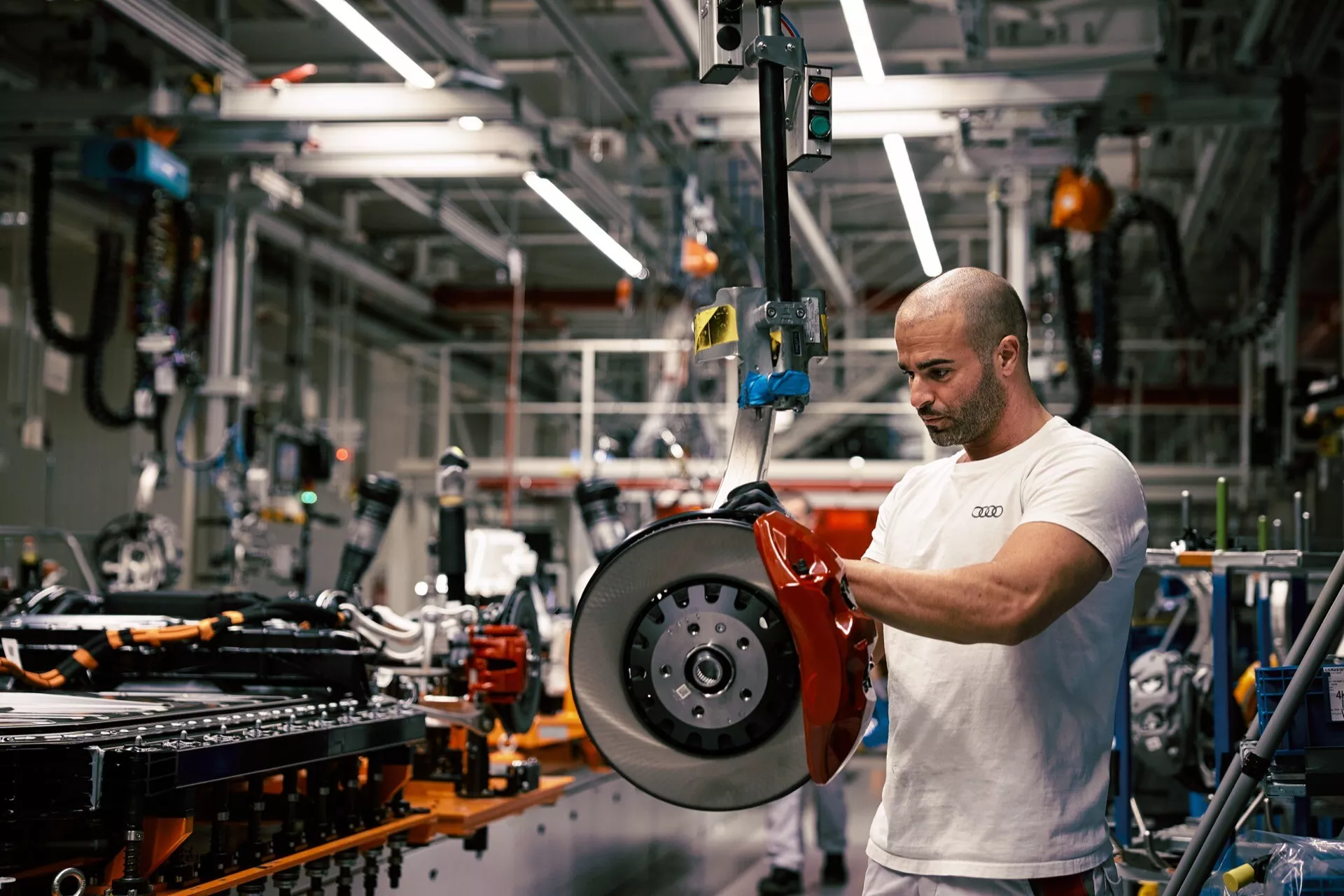 Zblízka pohľad na vysokovýkonnú červenú brzdovú strmene a ventilovaný brzdový kotúč v prostredí výroby automobilov, s technikom pracujúcim na montáži komponentu na šasi vozidla na pozadí