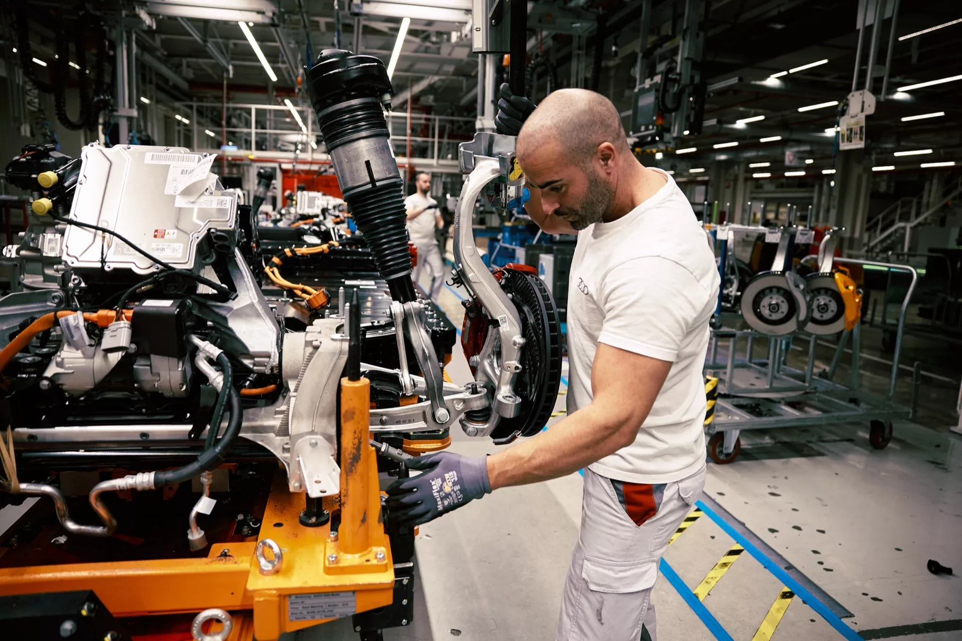 Zadný podvozok Audi Q8 e-tron 2023 na montážnom stupni s viditeľným zavesením a montážou brzdy, a pracovník používajúci elektrický nástroj vo výrobnej hale automobilov