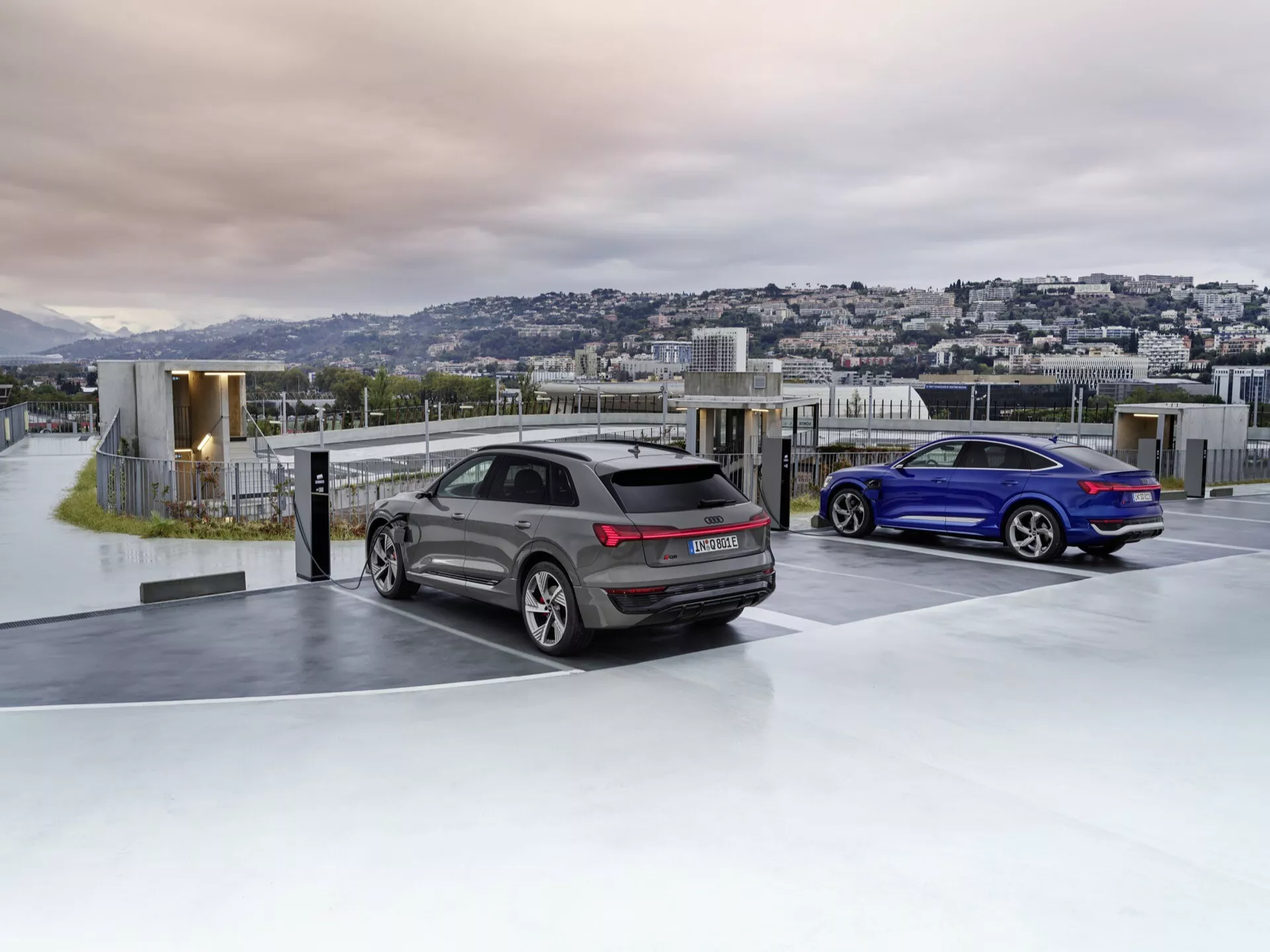 Audi Q8 e-tron 2023 v sivej farbe a športové kupé v modrej farbe zaparkované na luxusnom strešnom parkovisku na úsvite alebo súmraku s pozadím mestského pejzažu, predvádzajúce pokročilú aerodynamiku, športový dizajn, LED zadné svetlá, dvojitý výfuk a sklá pre súkromie