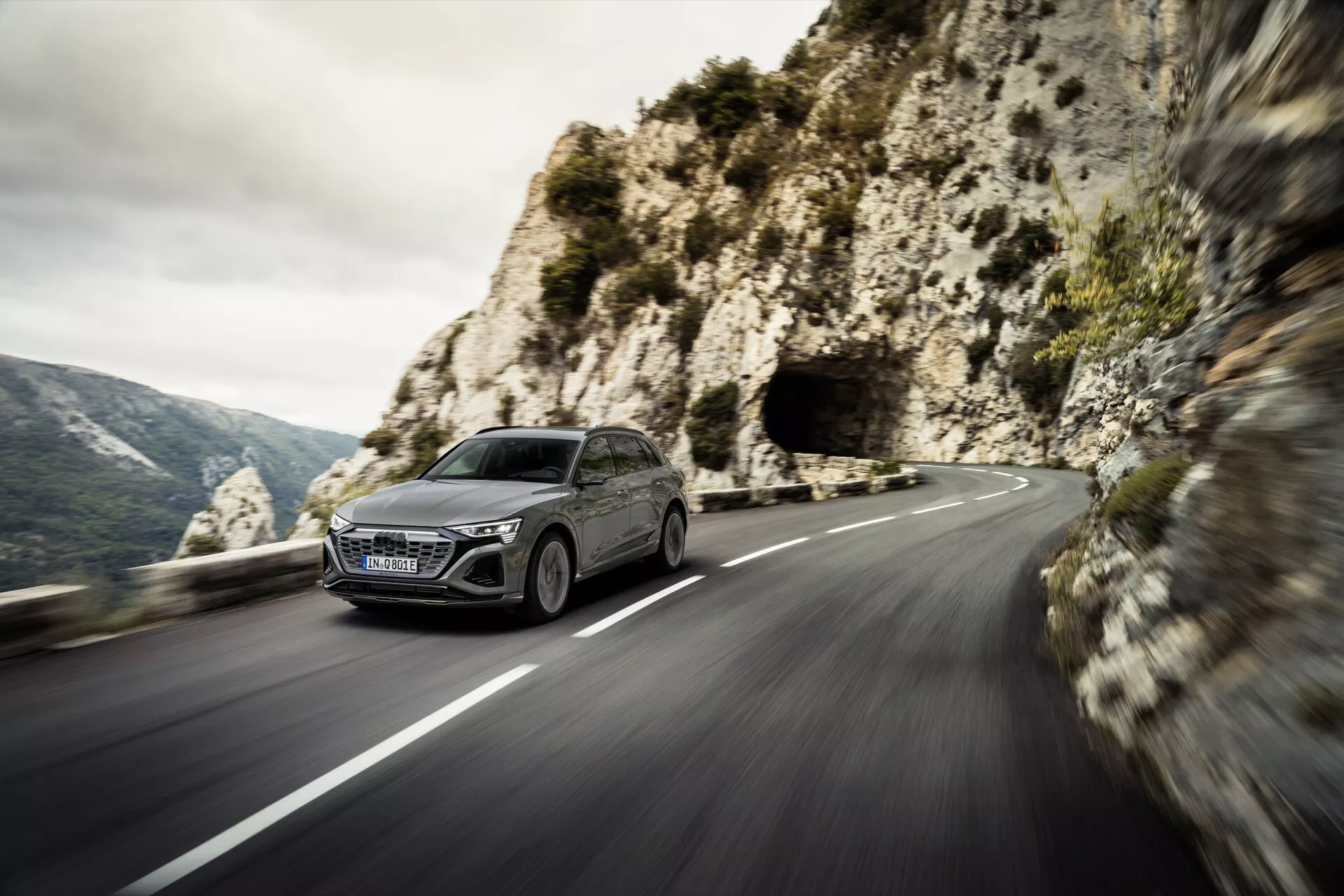 Audi Q8 e-tron 2023 dynamicky prechádza serpentínami na horskej ceste, predvádza moderný design hatchbacku, LED svetlomety a veľkú mriežku z pohľadu prednej štvrtiny s malebným horským pozadím