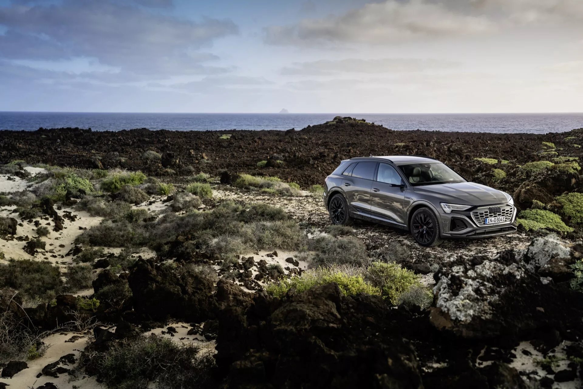 Audi Q8 e-tron 2023 v metalickej sivej farbe, predstavujúce luxus s elegantným, športovým dizajnom, zachytené z bočného predného uhla na nerovnom teréne pod jemným osvetlením, ukazujúce pokročilú technológiu LED svetlometov, výraznú mriežku s veľkým emblémom, dynamické kontúry boku a viaceré hovorové zliatinové kolesá.