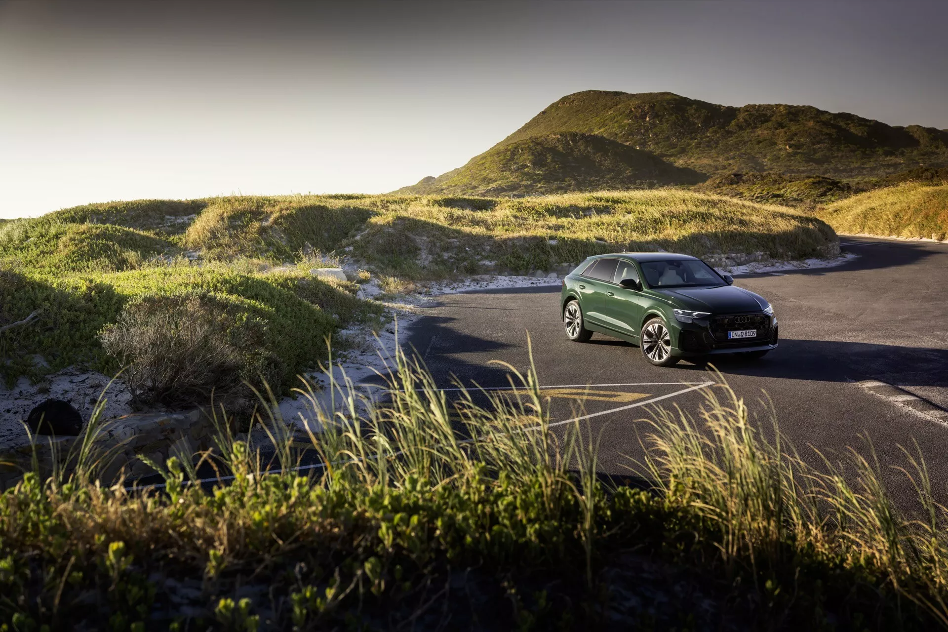 SUV Audi Q8 2023 (4M-facelift) zaparkované na pobrežnej ceste uprostred piesočných dún a trávy marram, osvetlené svetlom zlatej hodiny, ktoré zobrazuje prednú mriežku, súbor svetlometov a aerodynamický dizajn