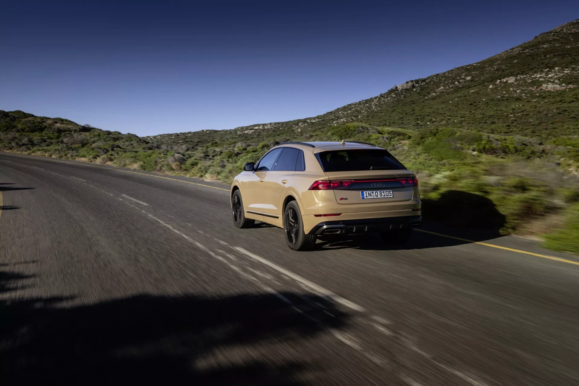Audi Q8 2023 (4M-Facelift) v pohybe, zachytené z zadného trojštvrťového uhla zobrazujúceho pravú stranu a zadnú časť na rozdelenej diaľnici s prirodzeným pozadím kopcov pod modrou oblohou, prezentujúce štýl strechy ako kupé, elegantný dizajn, predĺžené zadné svetlá, svetlo béžovú farbu a dvojitý výfukový systém.