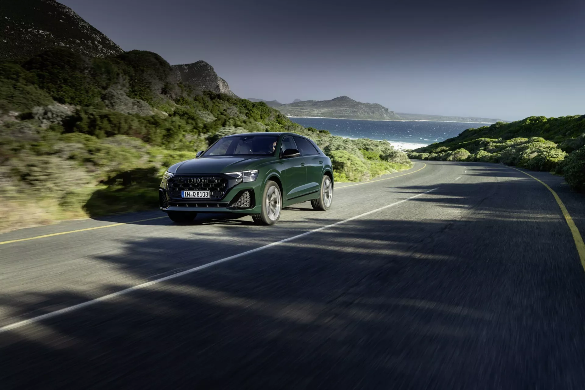 Audi Q8 2023 (4M-Facelift) idúce po pobrežnej ceste, zobrazujúce pohľad spredu a zboku na jeho veľkú mriežku, LED svetlomety, štíhlý kapot a čiastočný bok, zosobňujúce technológiu a luxus v pohybe na pozadí jasnej oblohy, hôr a oceánu.