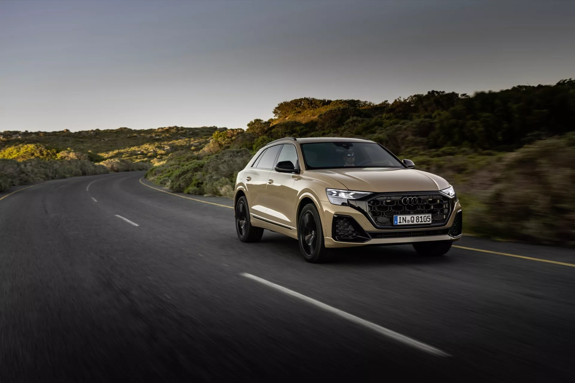 Audi Q8 2023 (4M-Facelift) v dynamickom pohybe po kľukatej ceste počas zlatého hodina, predstavuje športový dizajn kupé auta, agresívnu prednú časť s LED svetlometmi a šesťuholníkovou mriežkou a aerodynamický siluetu proti pozadí prírodnej krajiny.