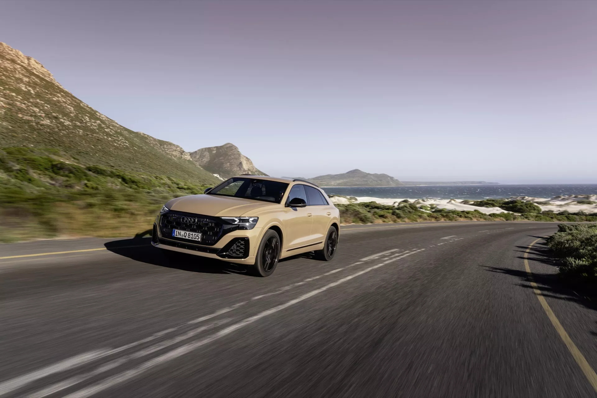 Trojštvrťový predný pohľad na Audi Q8 2023 (4M-Facelift) na pobrežnej ceste, predvádzajúci LED svetlomety, aerodynamický dizajn a rozostrenie pohybu naznačujúce pohyb