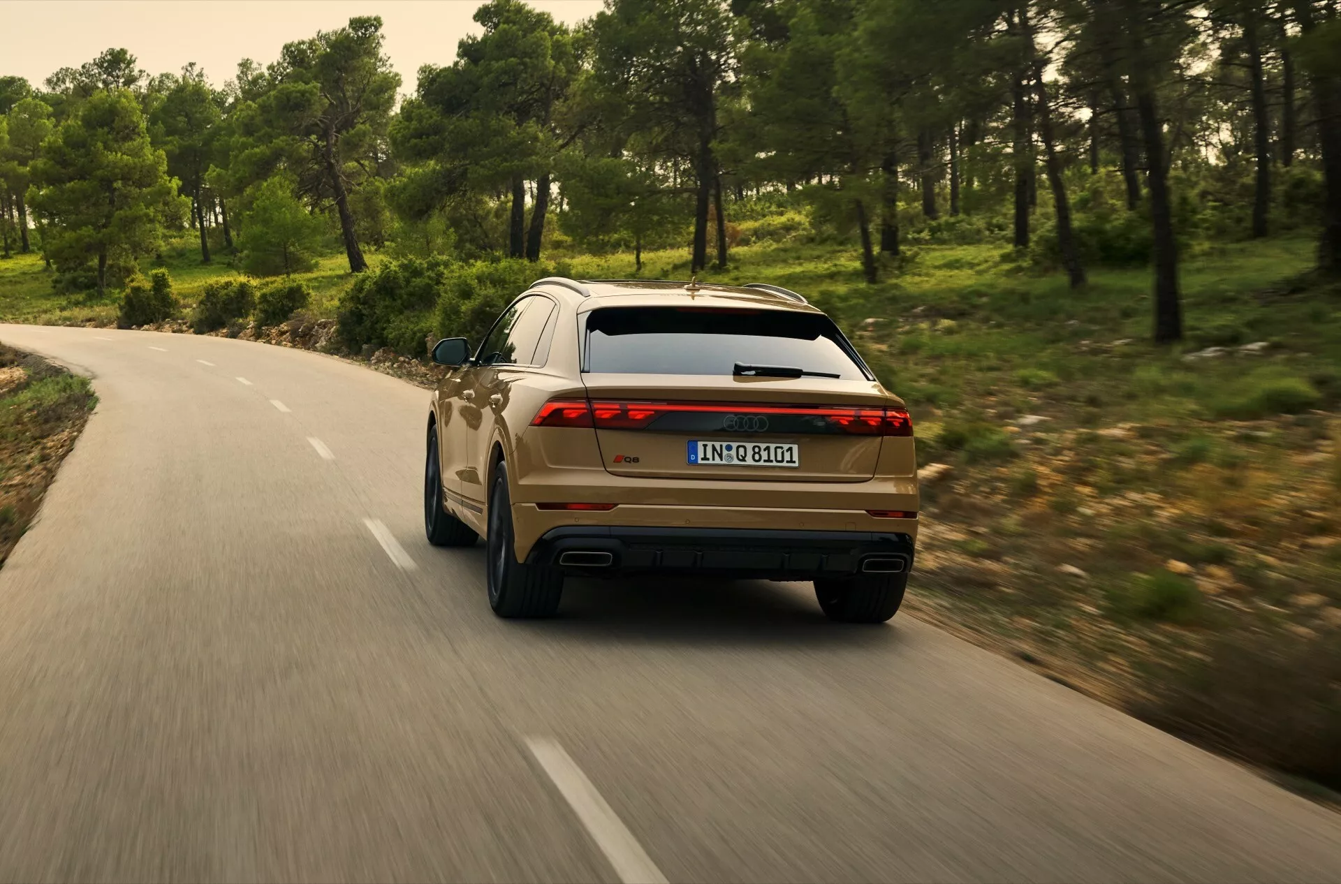 Zadný pohľad na Audi Q8 2023 (4M-Facelift) idúce po kľukatej ceste cez les, predvádzajúce pravý zadný štvrť s LED zadnými svetlami, dvojitým výfukom a modernými dizajnovými prvkami