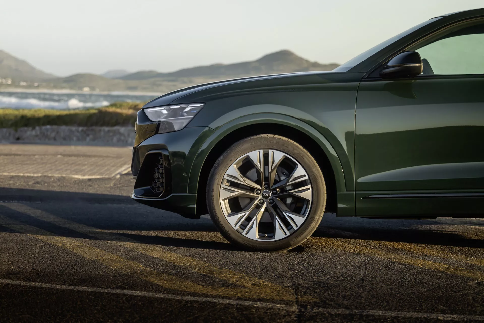 Detailný pohľad na športovú prednú ľavú štvrtinu Audi Q8 2023 (4M-Facelift), zahŕňajúci leštené viackolieskové zliatinové koleso, čiastočný predný nárazník s veľkým prívodom vzduchu, okraj moderne LED svetlomety a lesklý hlboko zelený metalický lak na karosérii, proti vonkajšiemu pozadiu.