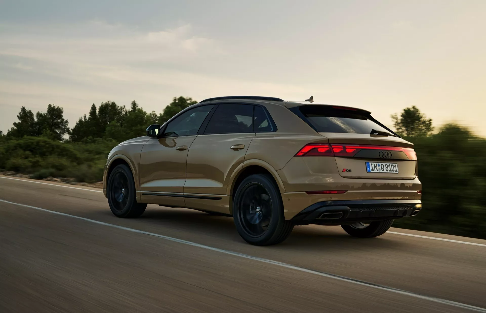 Zadný trojštvrťový pohľad na SUV Audi Q8 (4M-Facelift) 2023 v pohybe, s charakteristickým dizajnom zadných svetiel, kontúrovými líniami a profilom kolesa, zachytený počas dynamického jazdenia v vidieckom prostredí s teplým osvetlením.