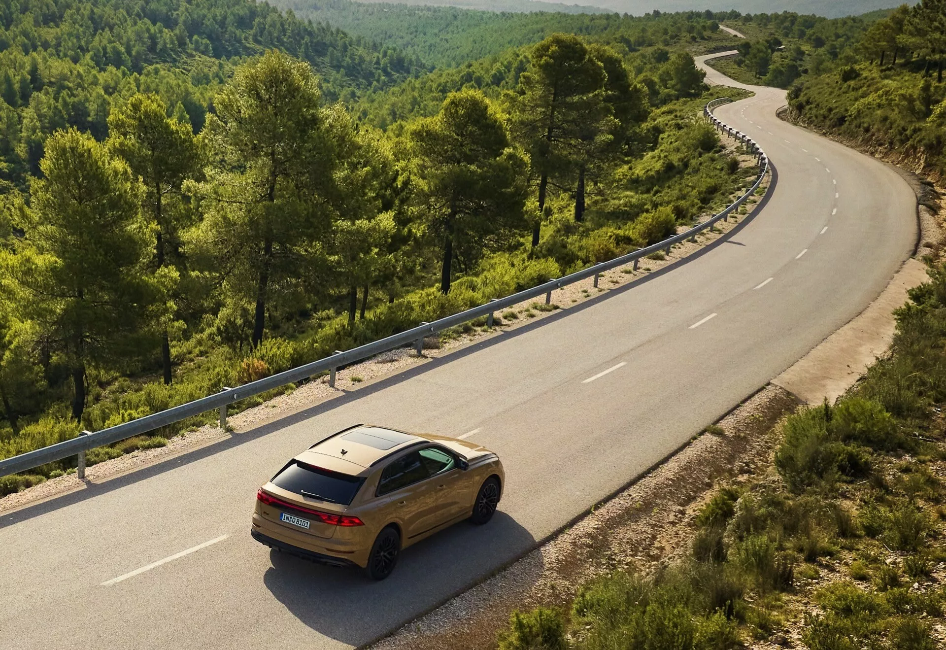 Zlatý Audi Q8 2023 (4M-Facelift) na kľukatej dvojprúdovej ceste v rurálnom alebo lesnom prostredí, pohľad z výšky ukazujúci jeho zadný a bočný dizajn proti prírodnej krajine