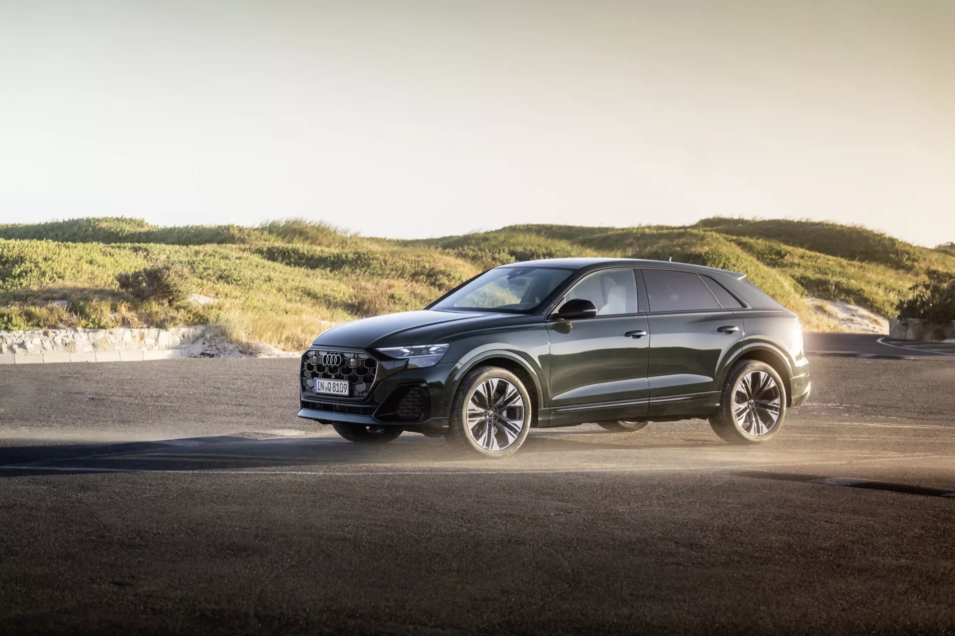 Moderný crossover Audi Q8 2023 (4M-Facelift) v exteriéri počas zlatej hodiny, zobrazujúci pokročilé LED svetlomety, luxusnú mriežku s bezpečnostnými funkciami, veľké zliatinové kolesá zamerané na výkon, metalický lak a športové dizajnové línie na pozadí asfaltu a mäkkých kopcov, zdôrazňujúce všestranné schopnosti a dizajn