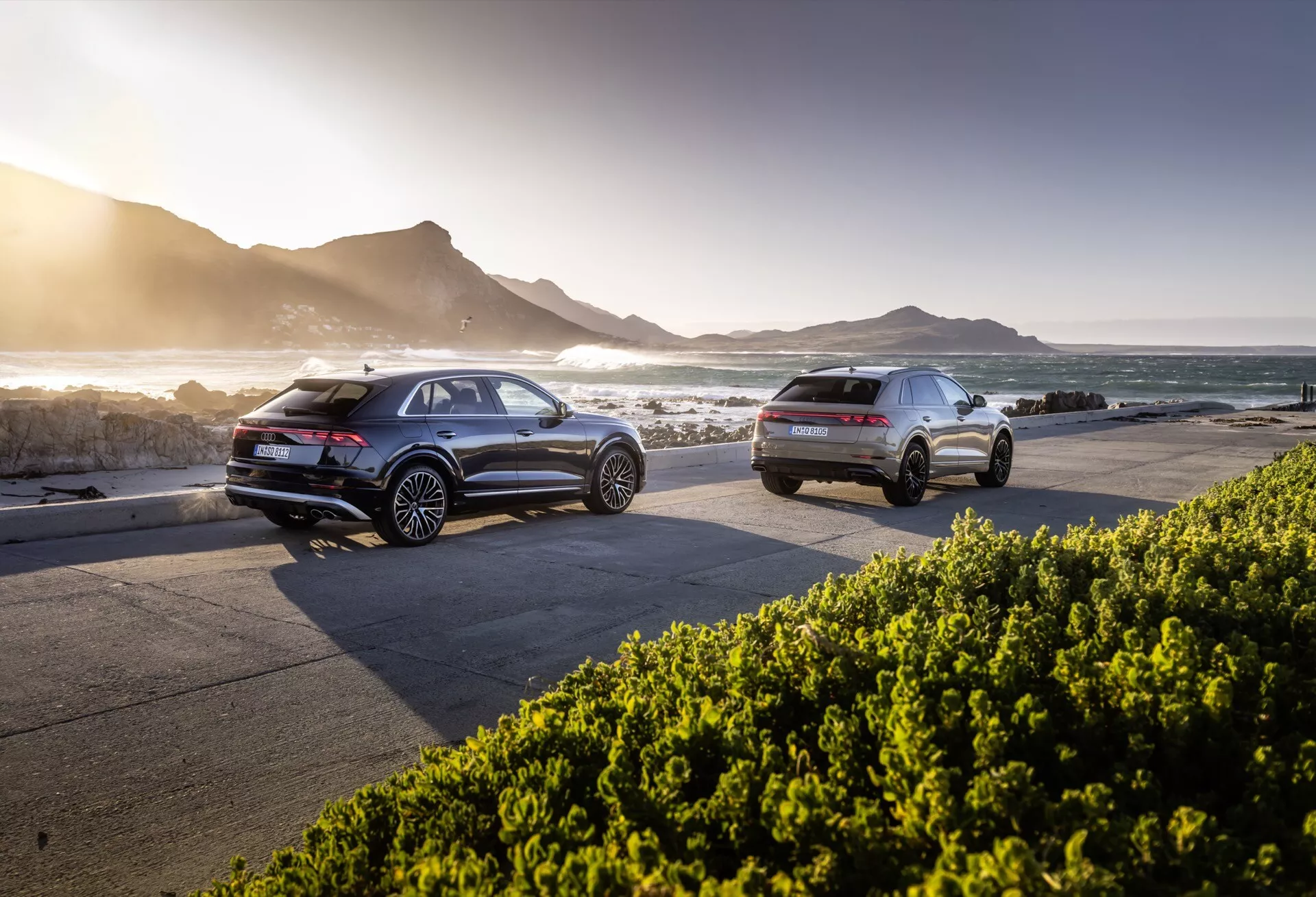 Modely Audi Q8 4M-Facelift z roku 2023 zaparkované vedľa seba na pobrežnej ceste pri západe slnka, s pohľadmi na zadný a bočný profil, pokročilou technológiou a malebným pozadím oceánu a hôr