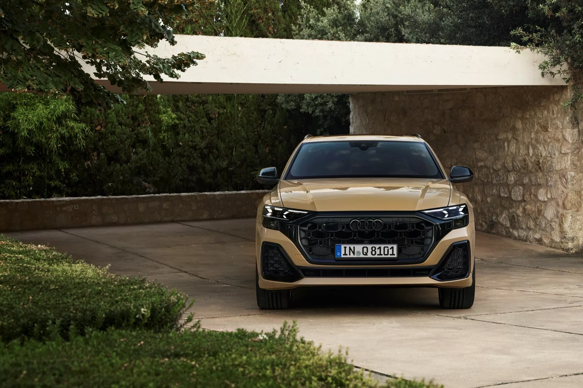 Audi Q8 2023 (4M-facelift) zobrazené z frontálneho uhla, zdôrazňujúce jeho širokú mriežku, štíhle LED svetlomety a metalický finiš, umiestnené na príjazdovej ceste obklopené bujnou zeleňou