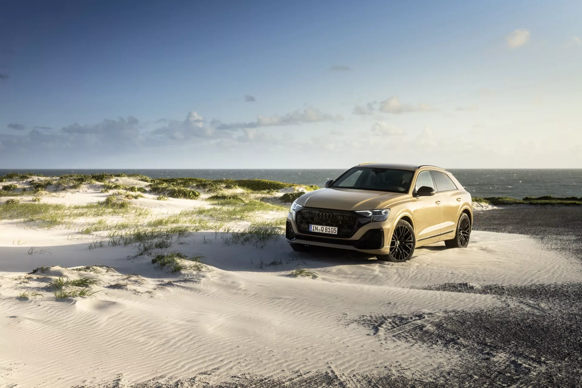 Audi Q8 2023 (4M-Facelift) zaparkované na piesočnatom teréne pod čiastočne zamračenou oblohou s morom na pozadí, predvádzajúce prednú mriežku, LED svetlomety a aerodynamický dizajn